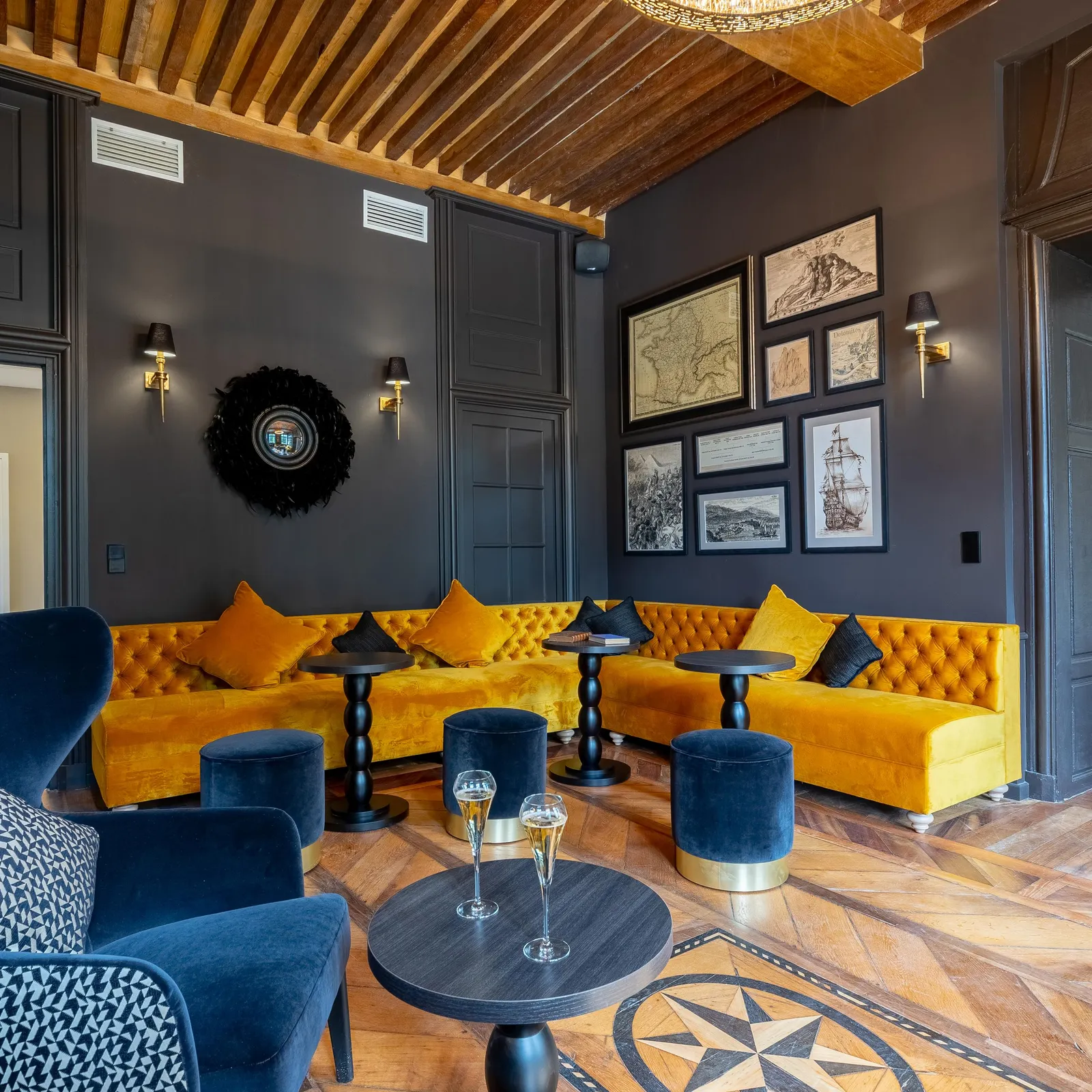 Un salon avec un canapé jaune, des fauteuils bleus, des tables basses et des décorations murales, sous un plafond en bois avec une suspension lumineuse