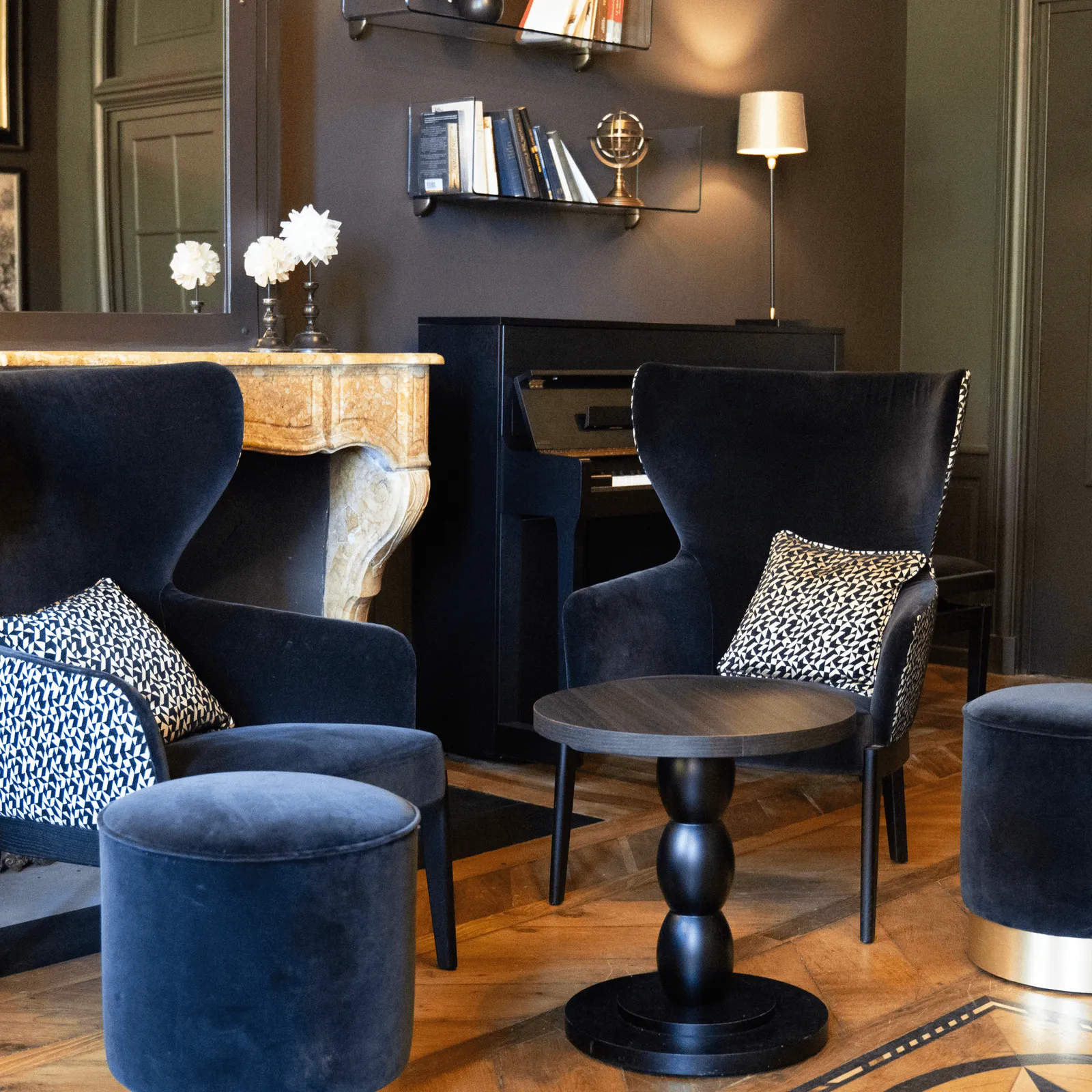 Salon avec des fauteuils et des ottomans en velours, une cheminée et des étagères avec des livres et des fleurs