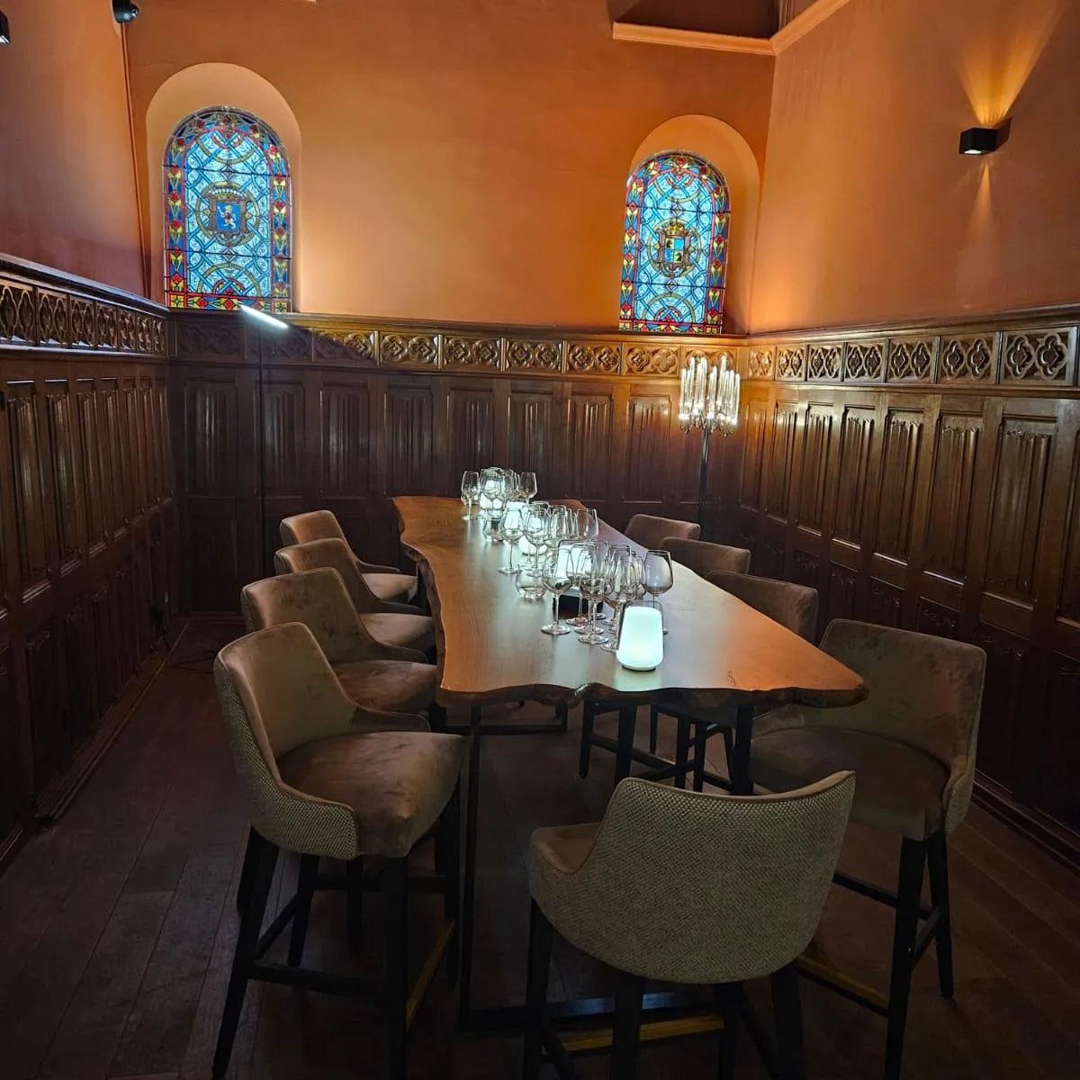A wooden table with glasses and candlesticks, surrounded by chairs, in a room with wooden walls and colored stained-glass windows.