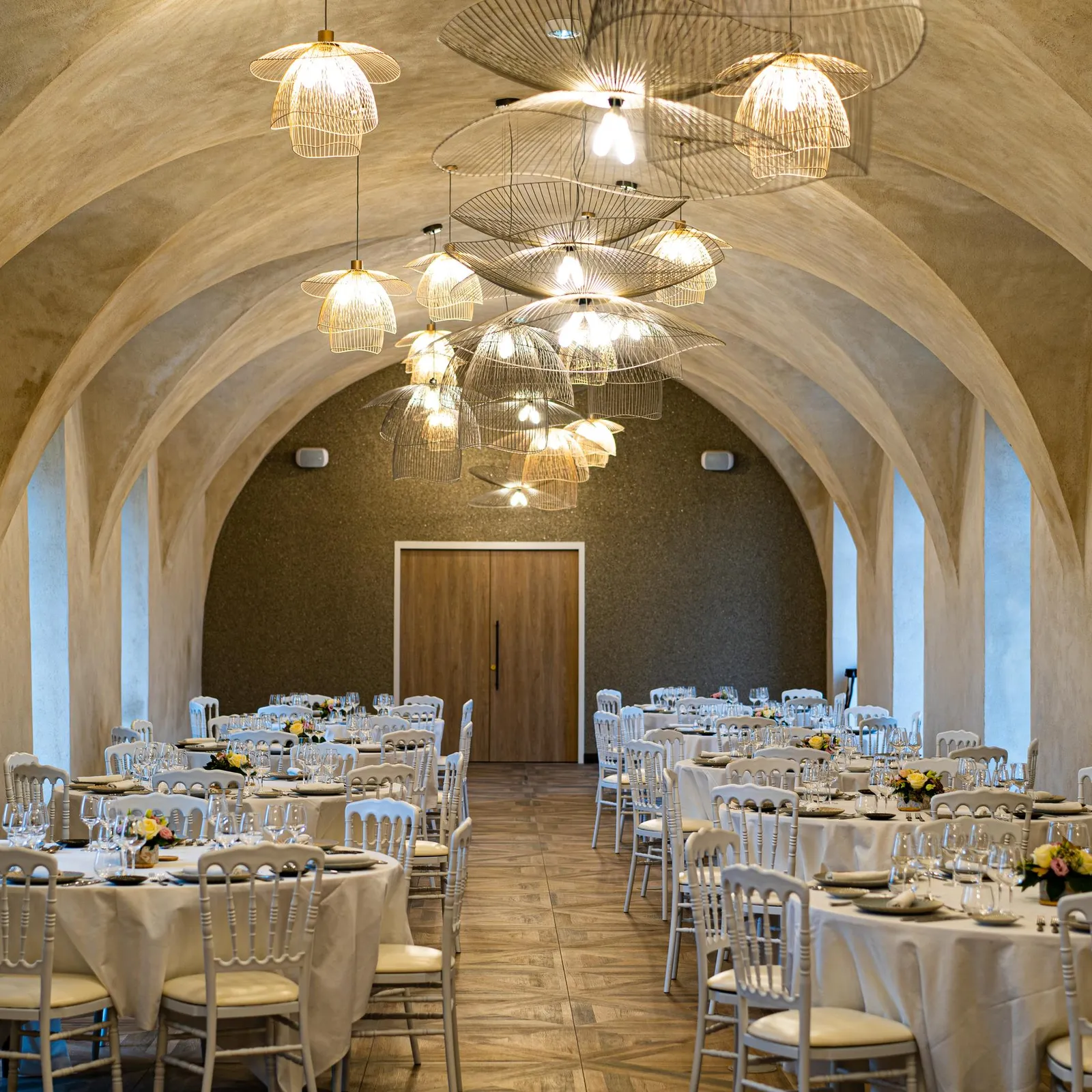 Salle de réception avec tables et chaises disposées pour un banquet, éclairée par des suspensions lumineuses au plafond