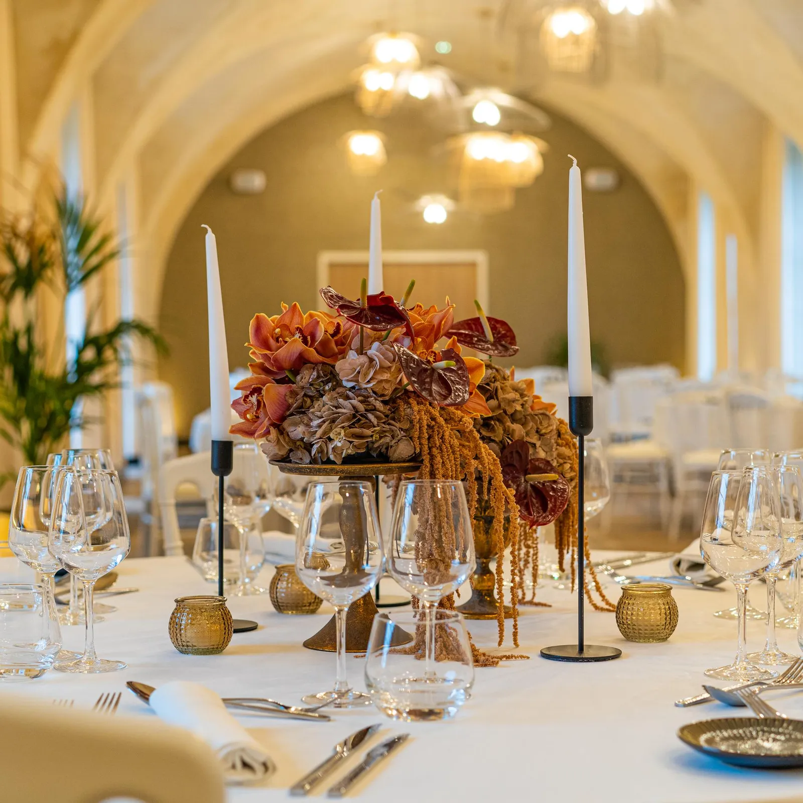 Table dressée avec verres, couverts, bougies et un centre de table floral dans une salle aux arches éclairées