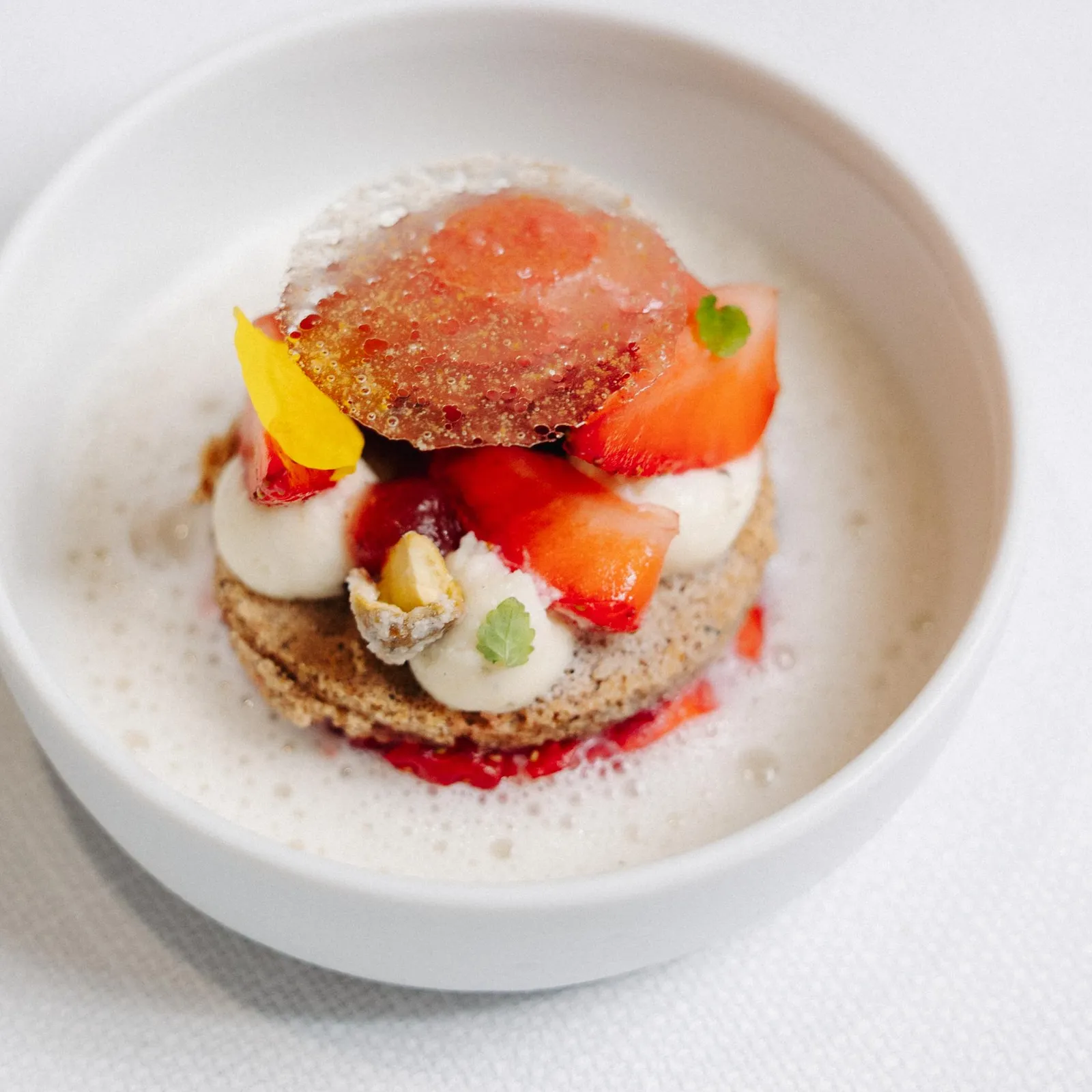 Un dessert dans un bol blanc composé d'une base de biscuit, de mousse, de morceaux de fraise et d'une fine tranche de fraise caramélisée