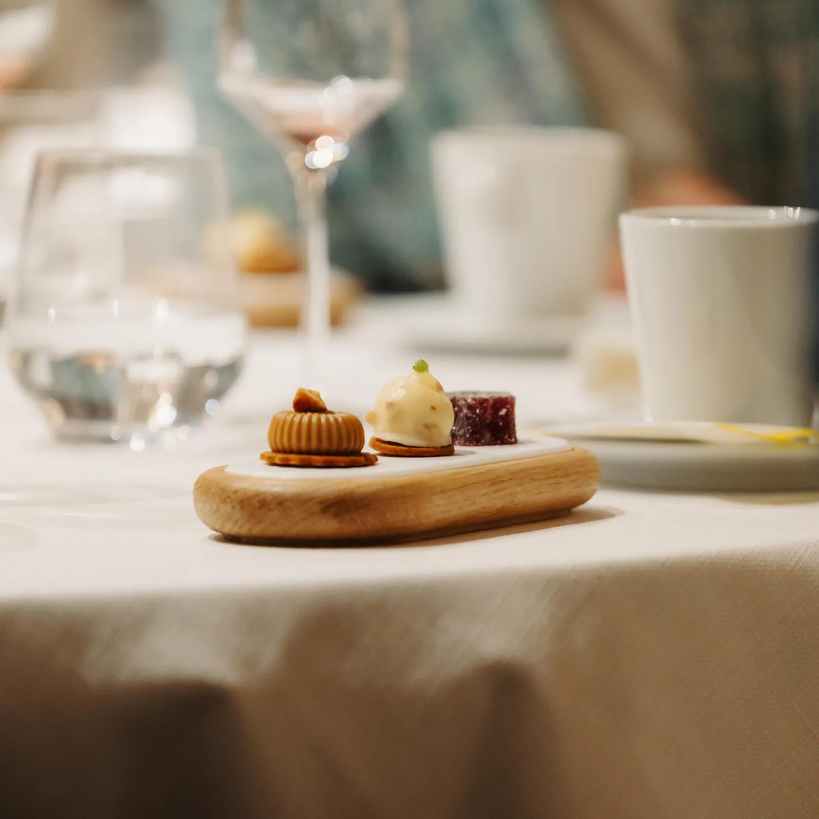 Petite assiette en bois avec trois desserts et verres de vin à l'arrière-plan sur une table recouverte d'une nappe blanche