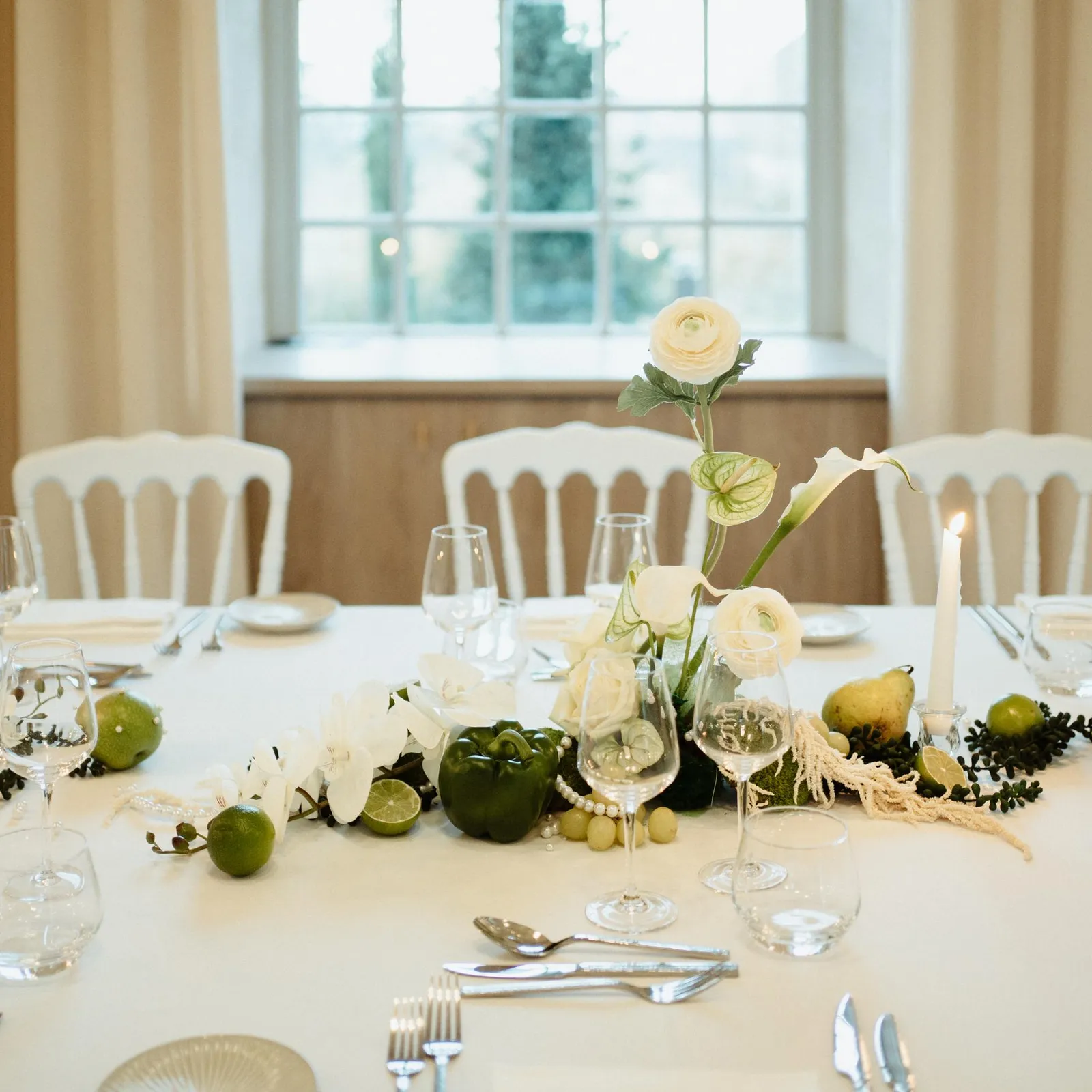Table dressée avec des verres, des couverts, des bougies et des fleurs blanches et vertes au centre, entourée de chaises blanches