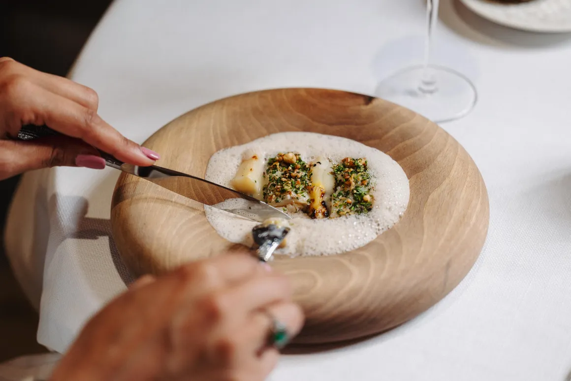Deux mains tiennent des couverts pour couper un plat gastronomique sur une assiette en bois posée sur une nappe blanche