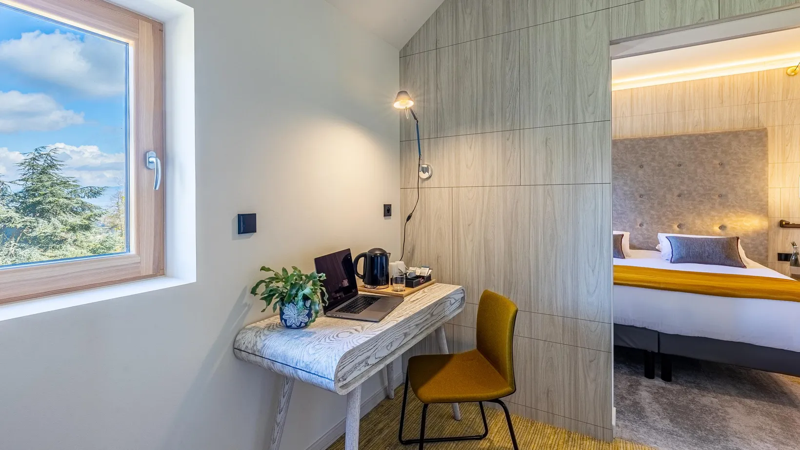 Bureau avec ordinateur portable, plante et bouilloire devant une fenêtre ouverte sur un paysage arboré, chambre avec lit visible en arrière-plan