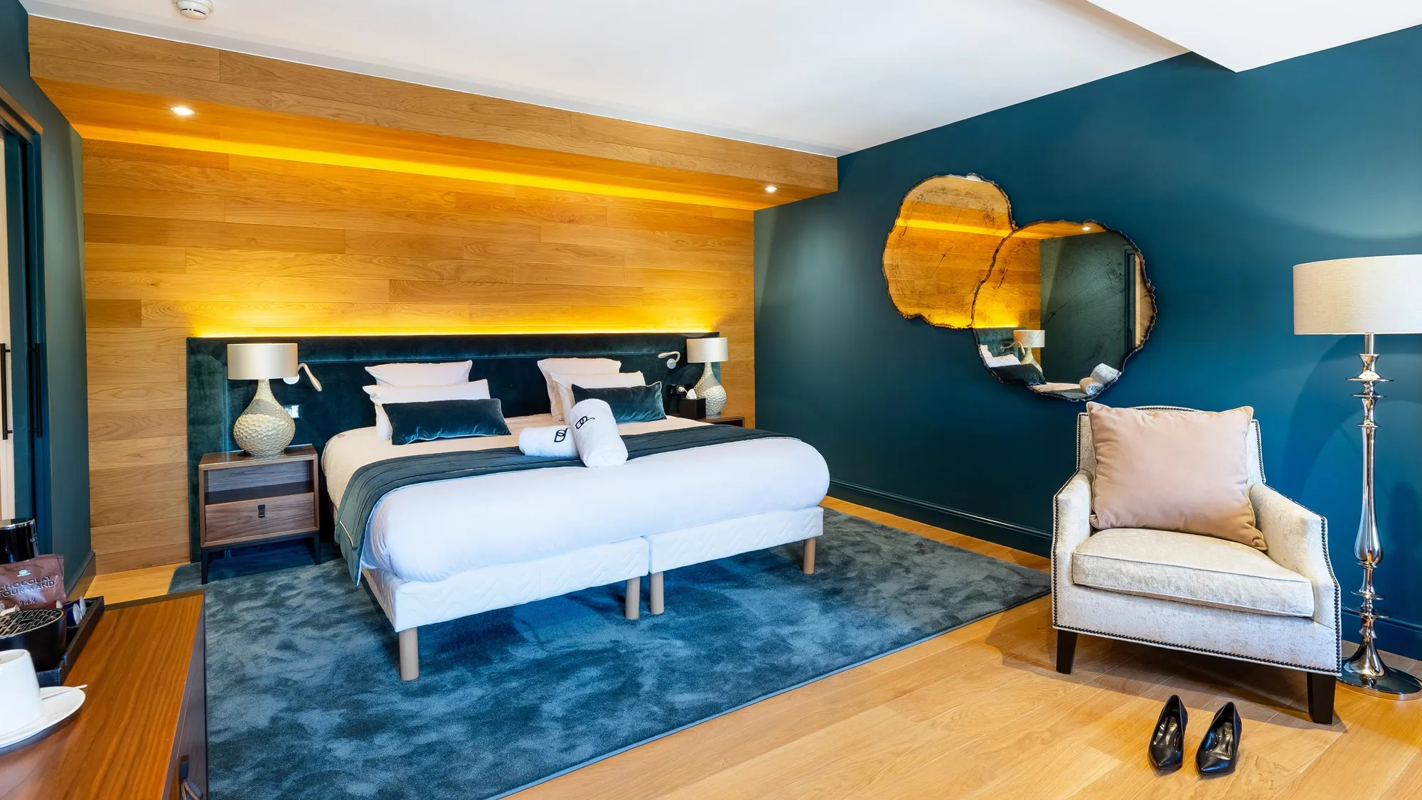 Room with double bed, round mirror, armchair and bedside table, dark blue walls and light wood, soft lighting
