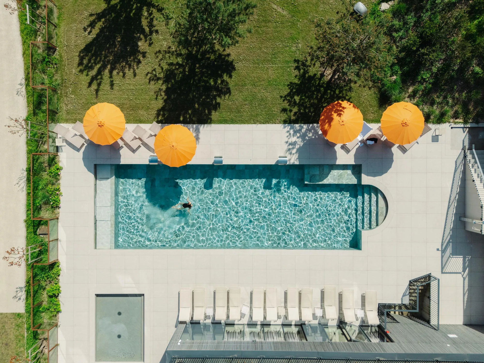 Vue aérienne d'une piscine rectangulaire avec une personne nageant, entourée de chaises longues et de parasols orange, dans un jardin avec pelouse et arbres