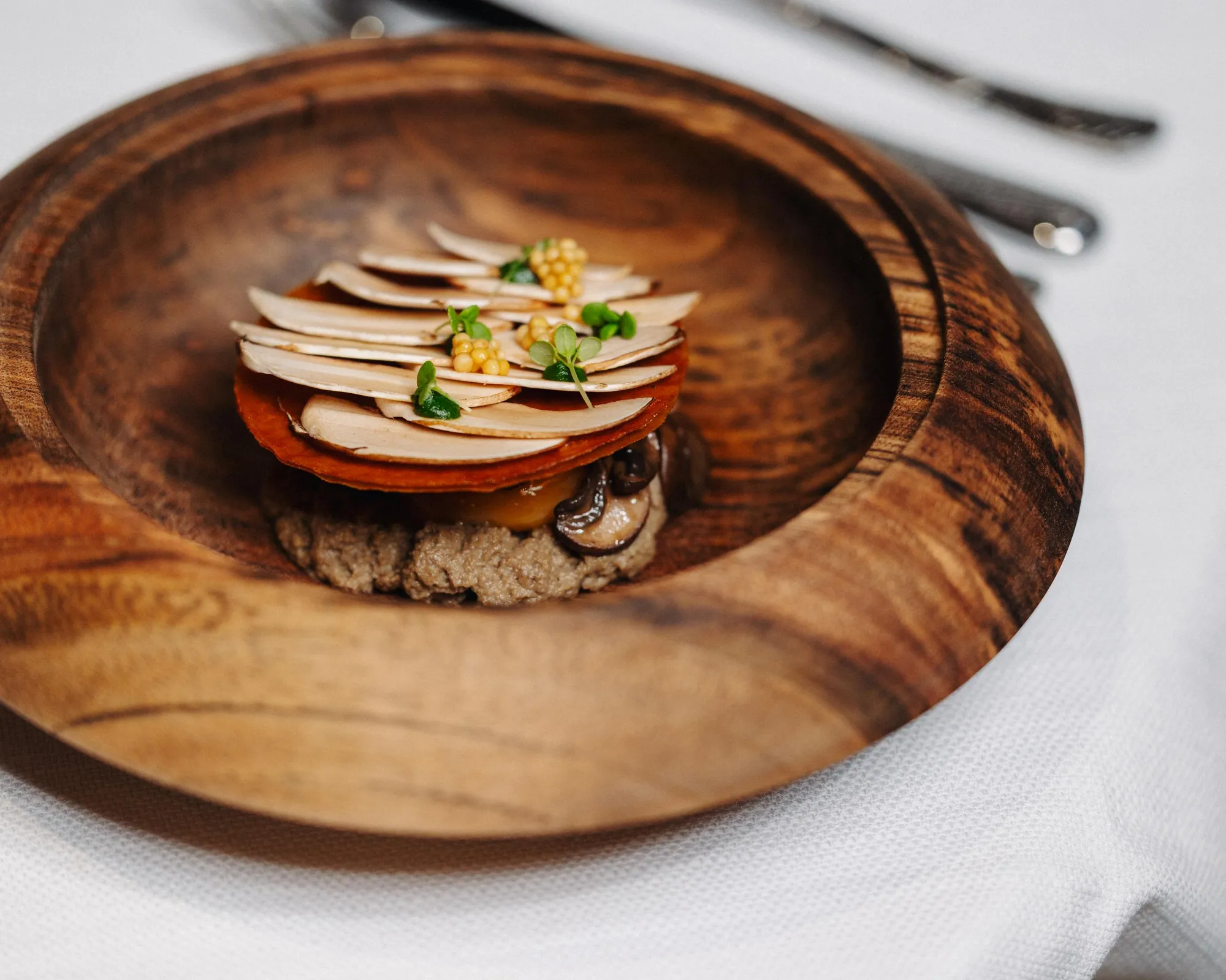Un plat en bois contenant une pile de fines tranches de légumes, de champignons et de graines, posé sur une nappe blanche
