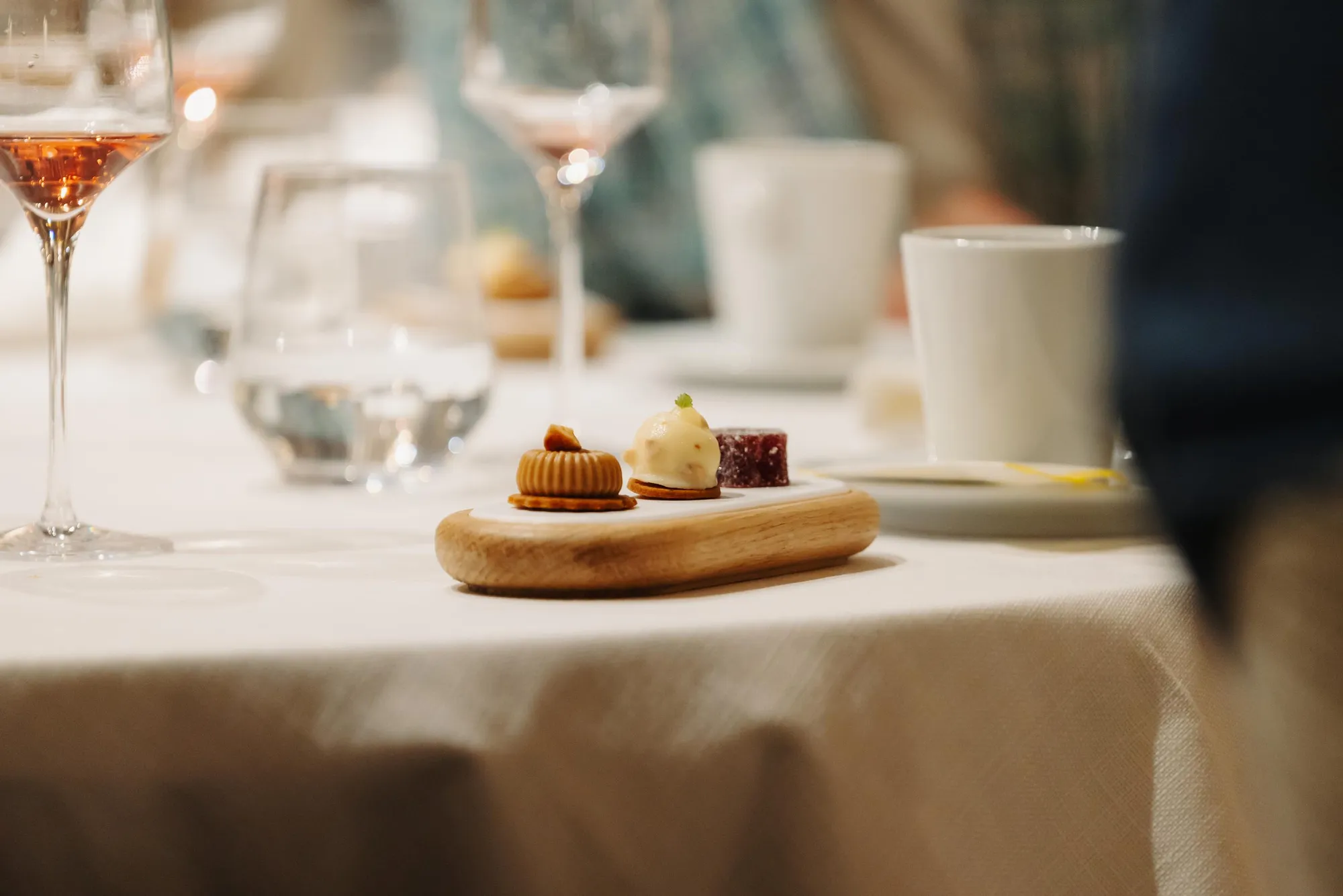 Small wooden plate with three desserts and wine glasses in the background on a table covered with a white tablecloth