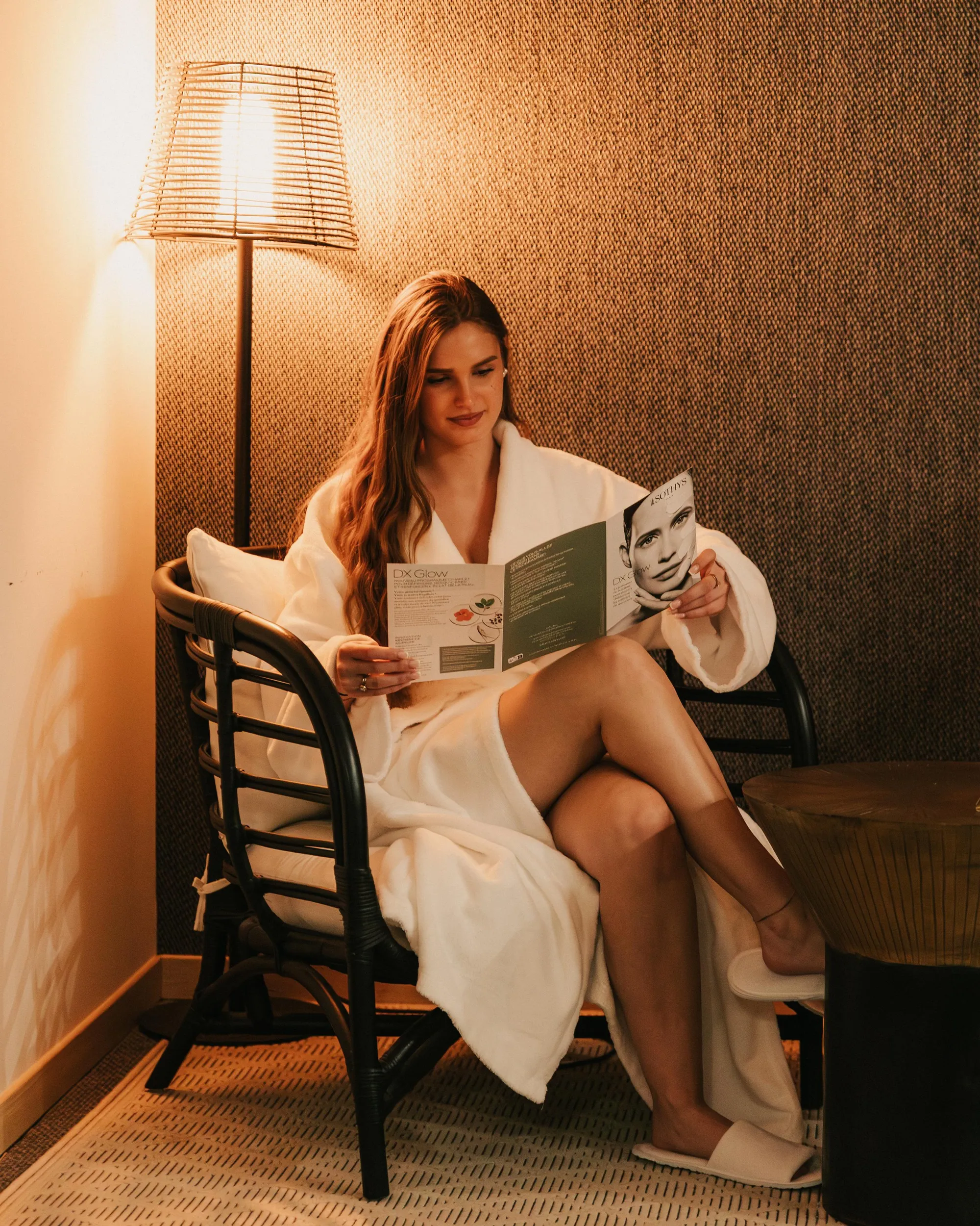 Une femme en robe de bain lit un magazine dans un fauteuil près d'une lampe allumée