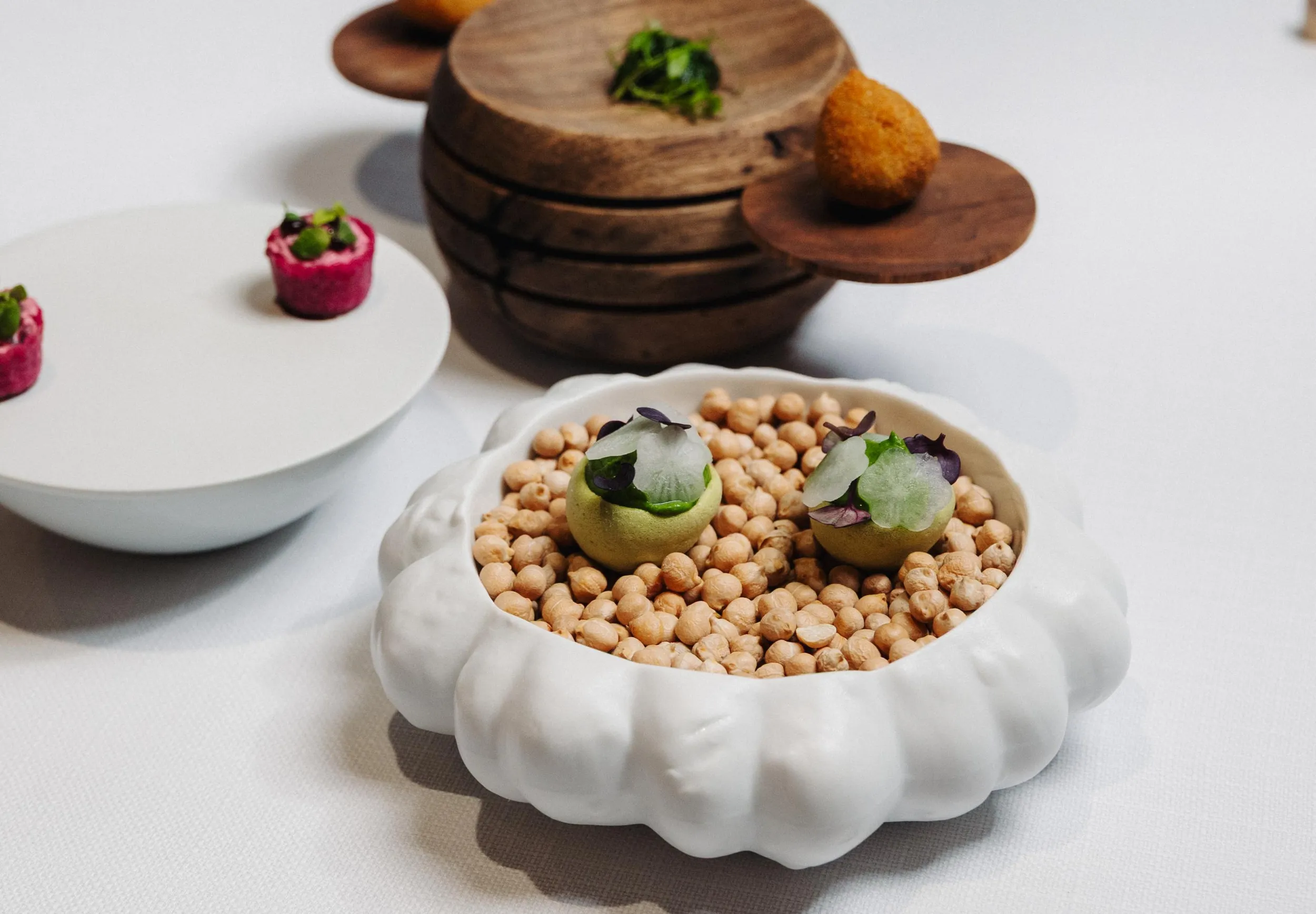 A white dish filled with chickpeas, topped with two green balls garnished with herbs and vegetable slices, placed on a white table.