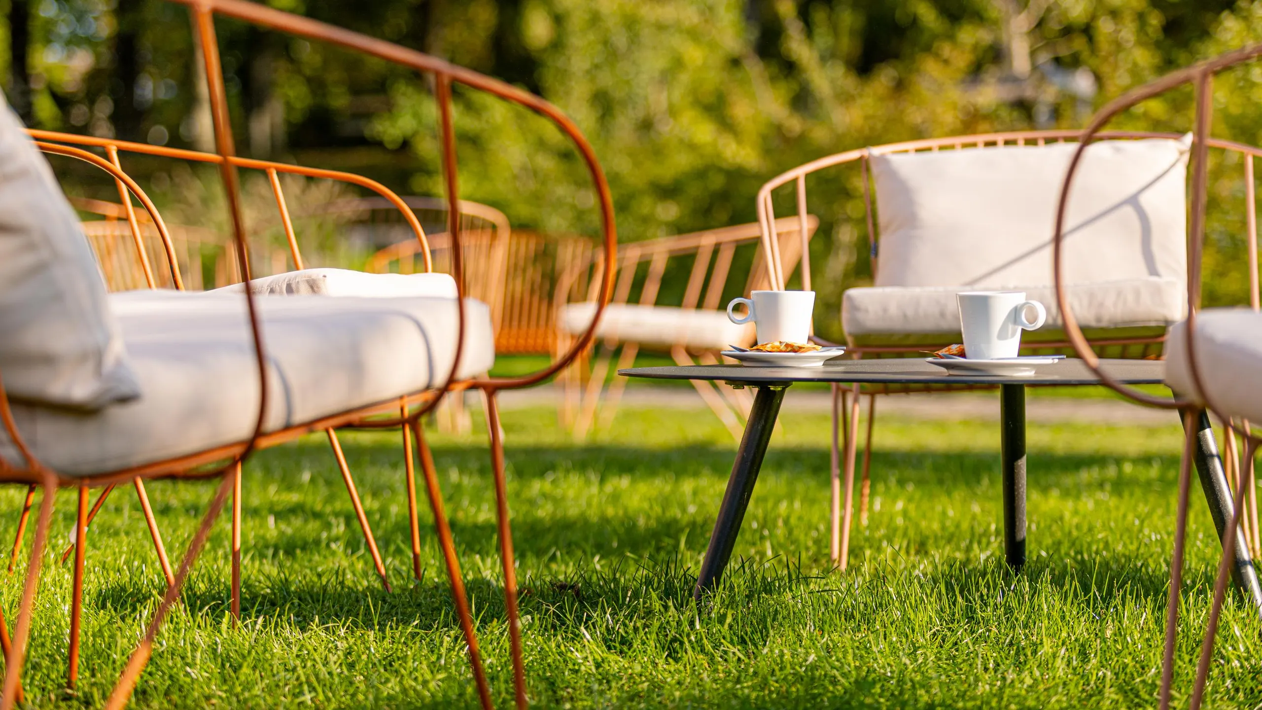 Quatre chaises en métal avec des coussins blancs et une table basse avec des tasses et des assiettes posées dessus sur une pelouse verte