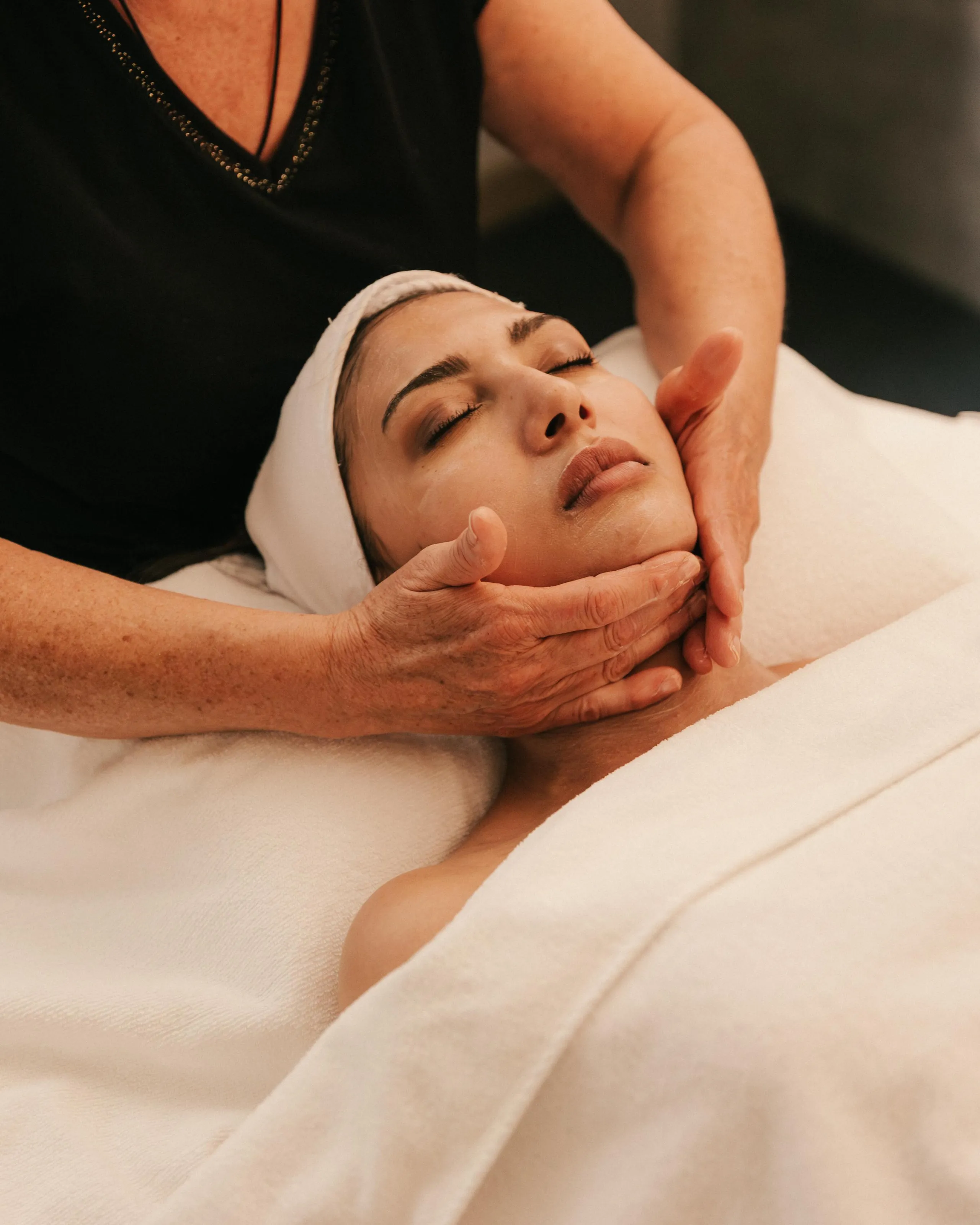 Une personne reçoit un soin du visage allongée sur une serviette blanche, les yeux fermés, tandis qu'une autre personne lui masse le visage