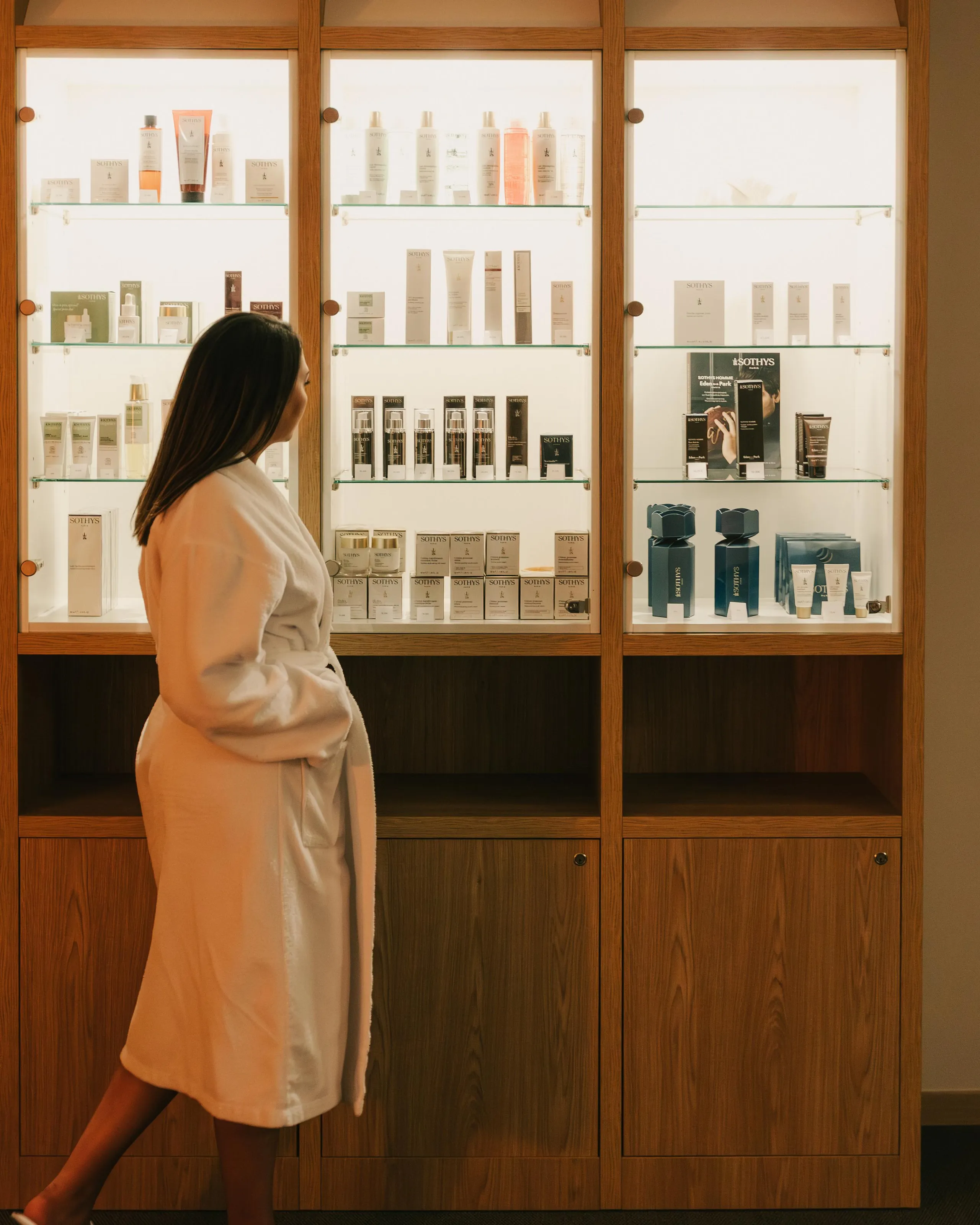 Une personne en robe de bain blanche marche devant des étagères vitrées remplies de produits cosmétiques Sothys