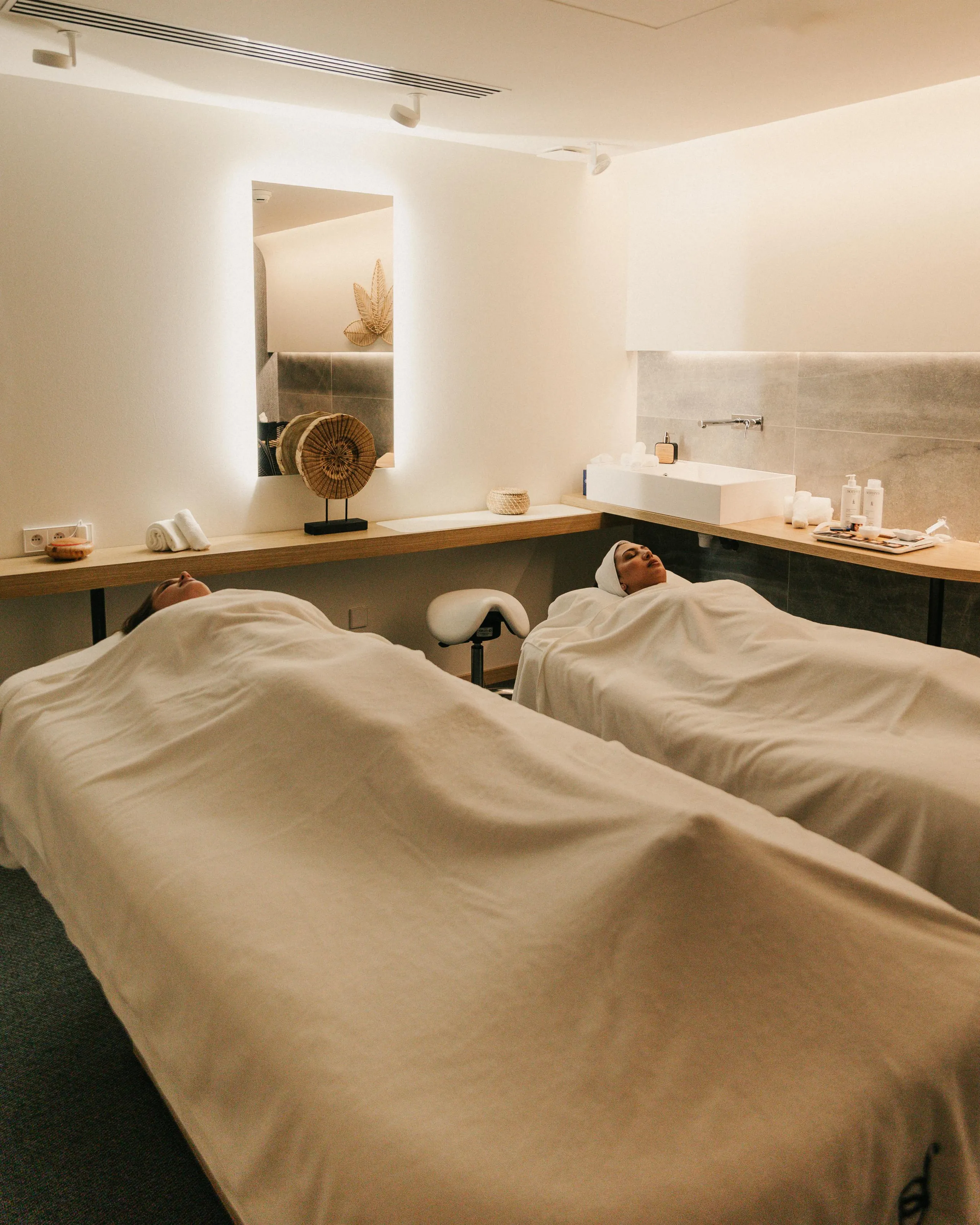 Deux personnes allongées sur des tables de massage dans une salle de soins, couvertes de draps blancs, avec des serviettes et des produits disposés sur une étagère
