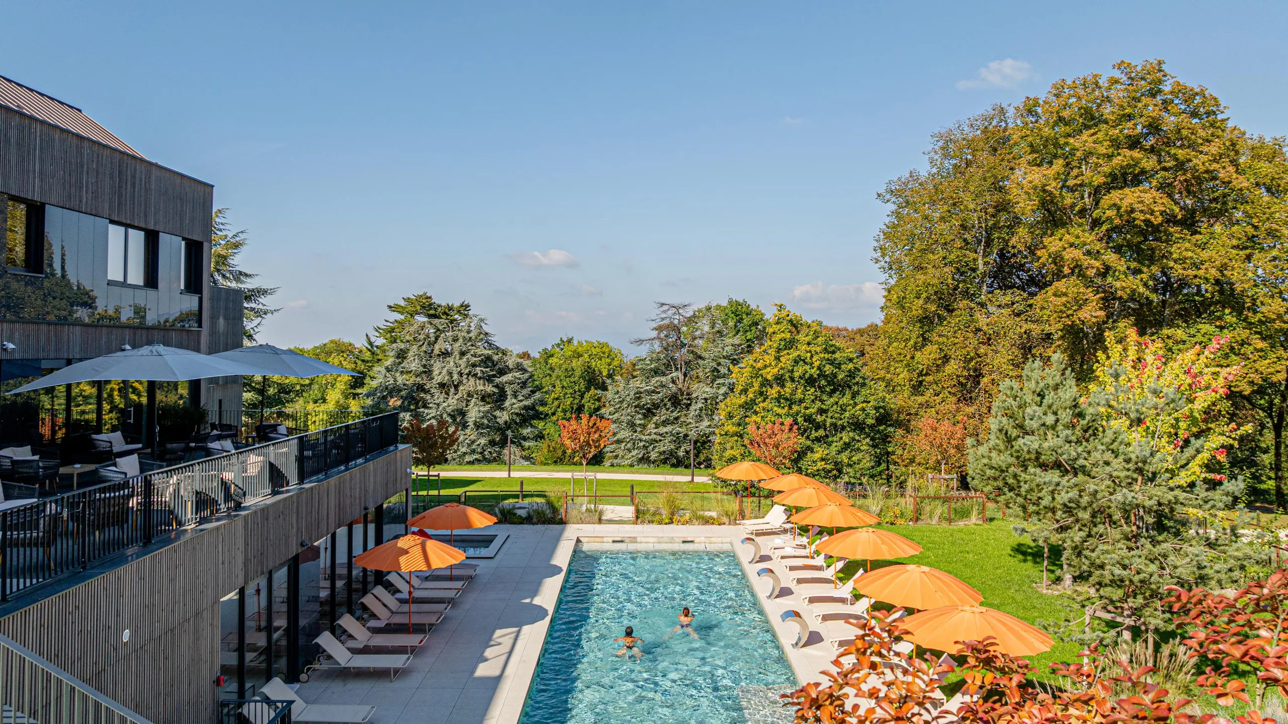 Piscine extérieure entourée de transats et de parasols, avec un bâtiment et un jardin arboré en arrière-plan