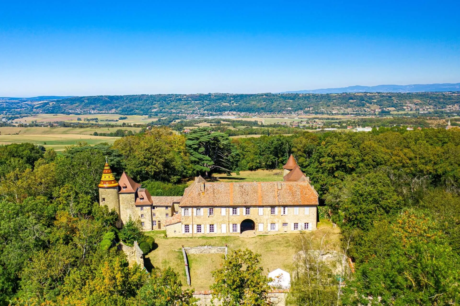 Ancien château entouré d'arbres avec toits en tuiles rouges et une tour à toit coloré, situé dans un paysage rural vallonné