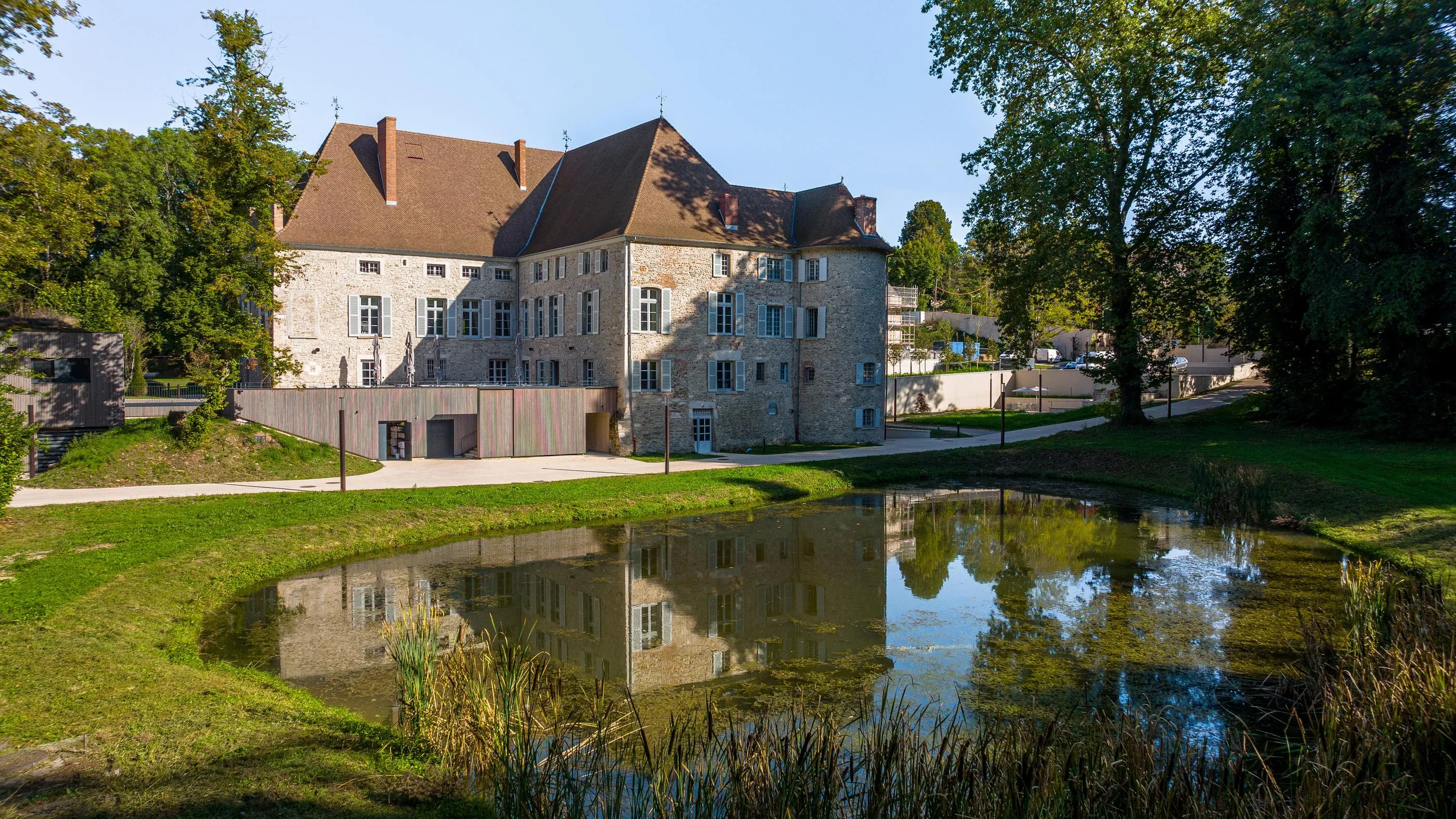 Ancien bâtiment en pierre avec toits en tuiles rouges entouré d'arbres, reflété dans un étang entouré d'herbe et de végétation