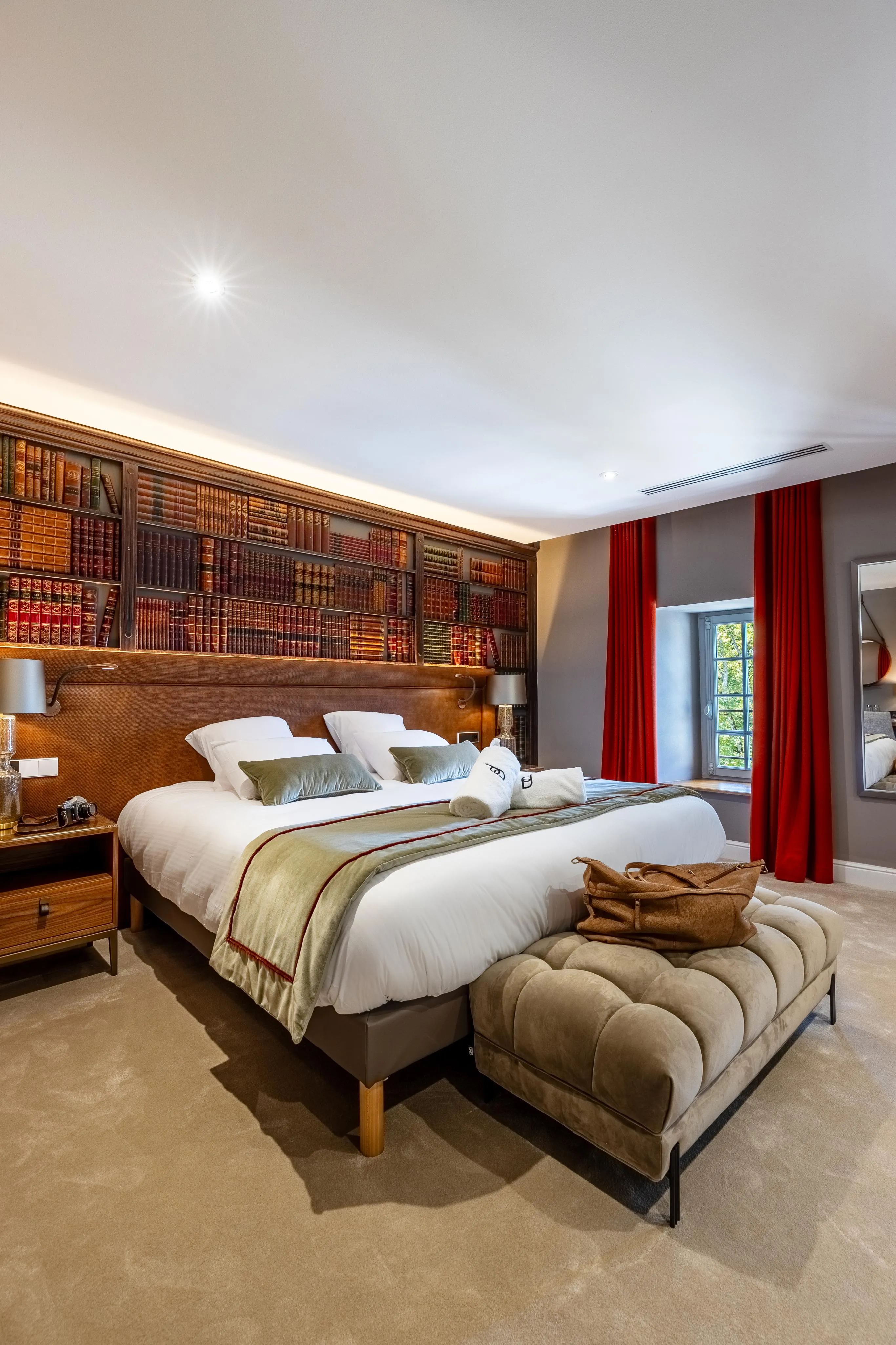 Chambre avec un grand lit, une tête de lit en bois avec étagères remplies de livres, un ottoman au pied du lit et des rideaux rouges