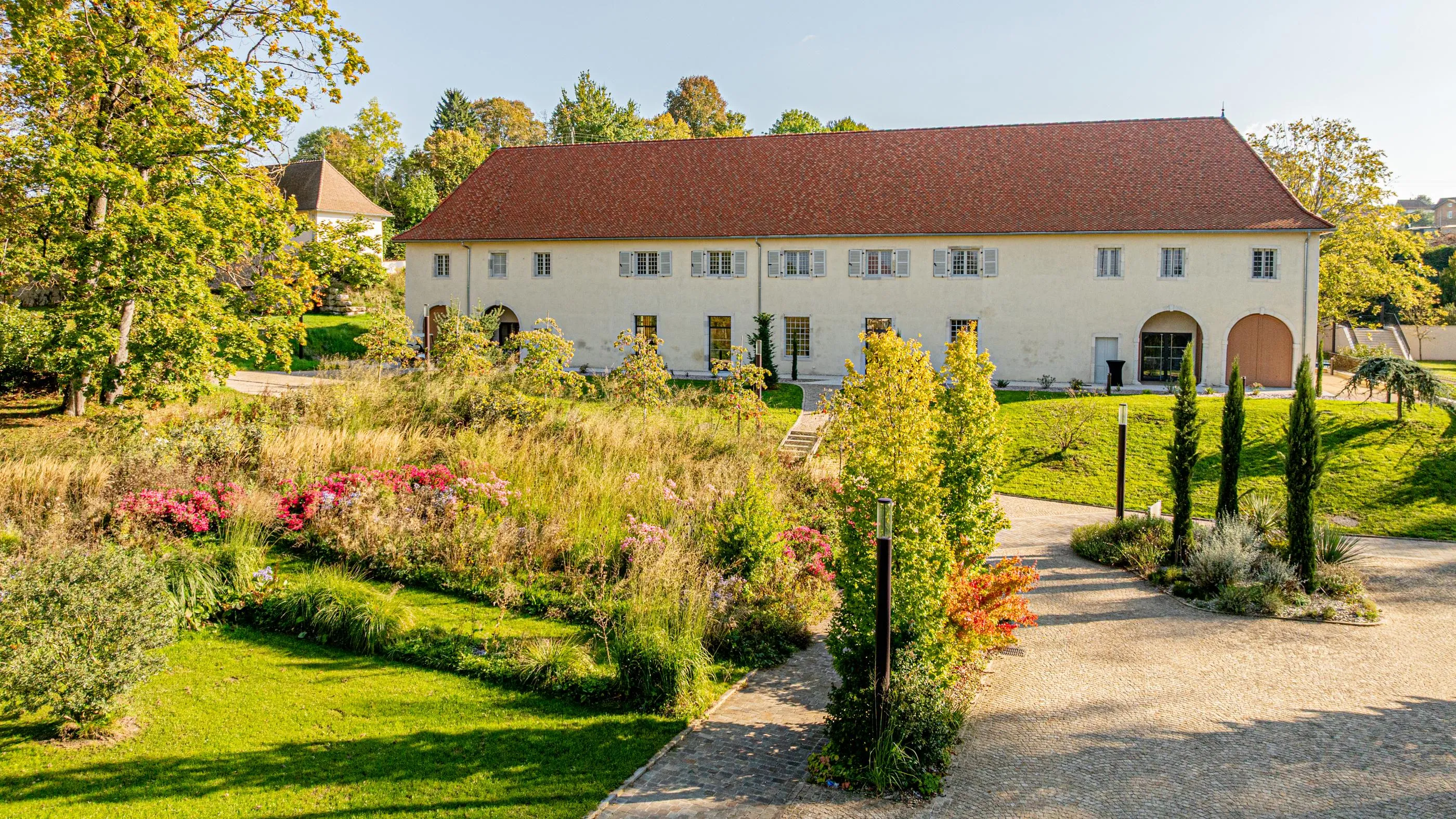 Un grand bâtiment à deux étages avec un toit rouge et des fenêtres blanches entouré d'un jardin bien entretenu avec des plantes et des arbres