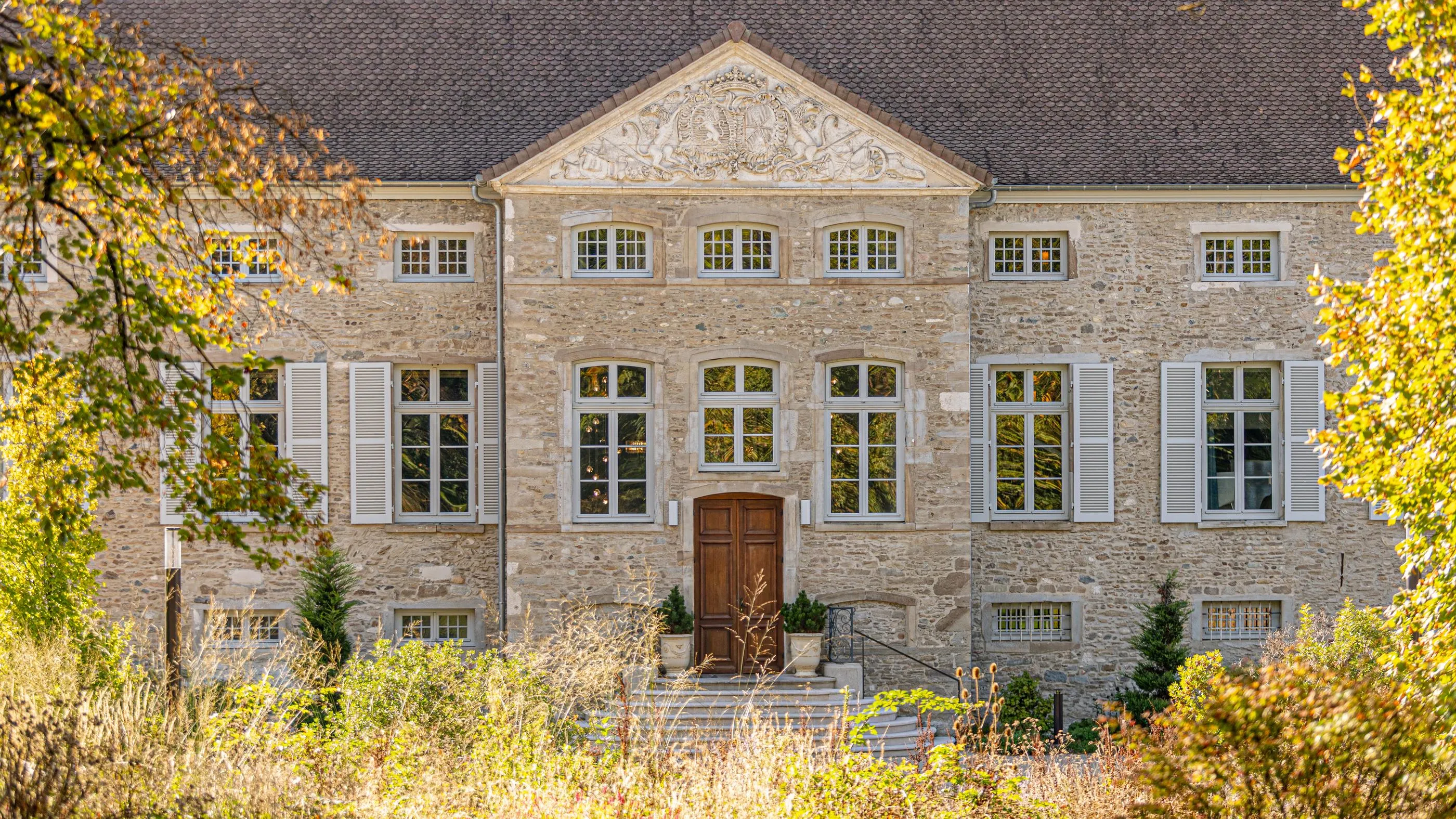 Ancienne maison en pierre avec des fenêtres à volets gris et une porte en bois, entourée de végétation automnale