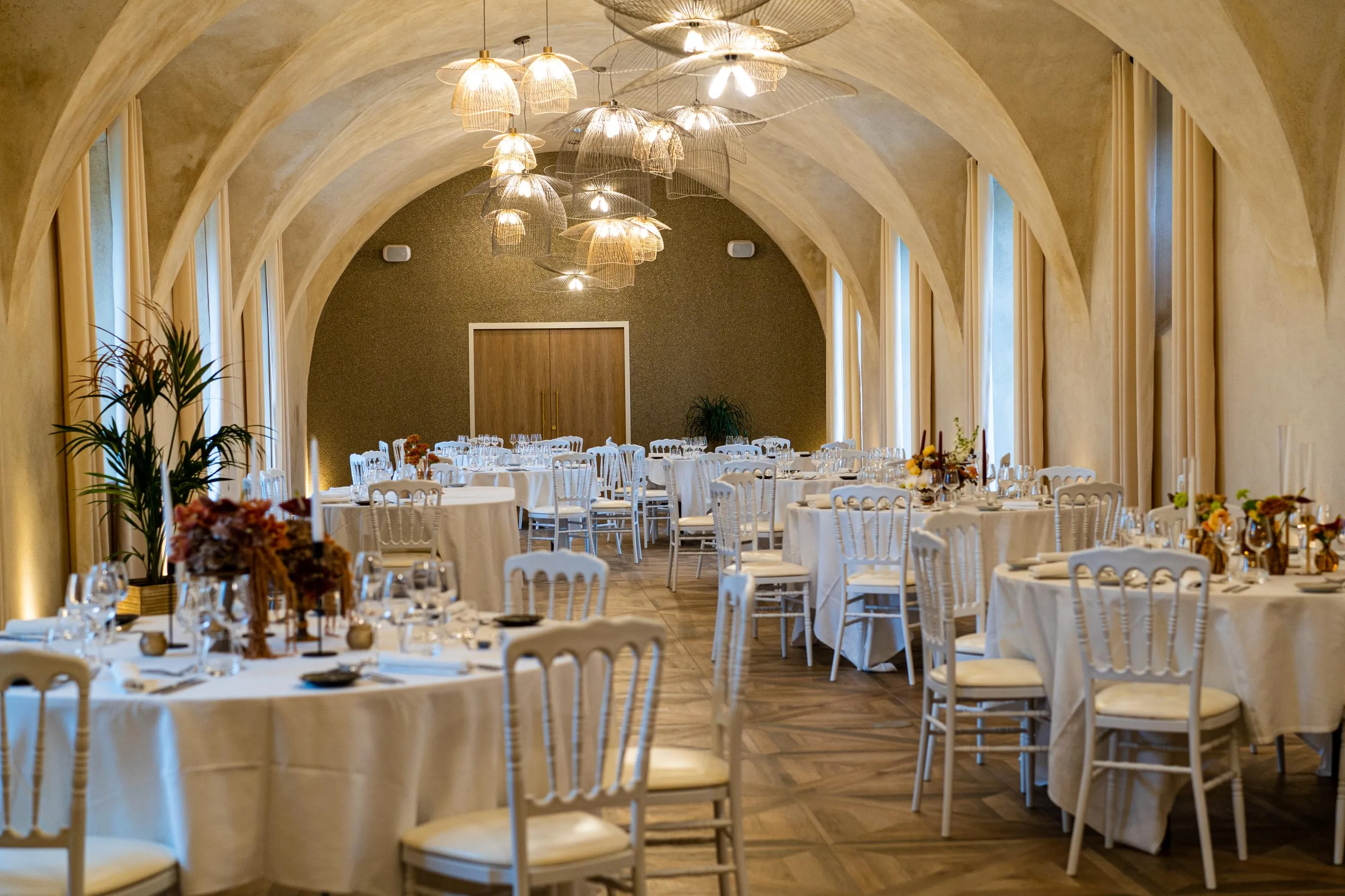 Salle de réception avec tables rondes recouvertes de nappes blanches, des chaises assorties et des décorations florales, éclairée par des suspensions lumineuses