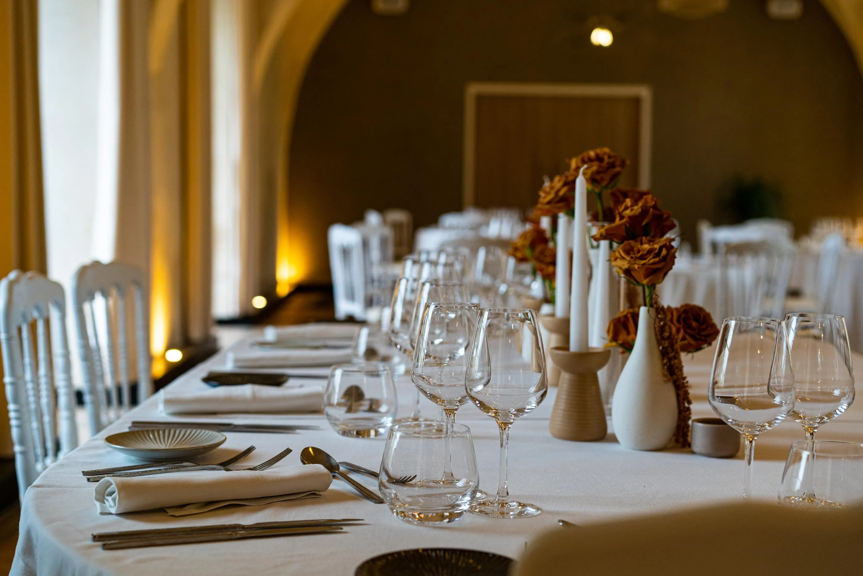 Table dressée avec verres, couverts, serviettes et bougies, décorée de vases contenant des fleurs, dans une salle avec des chaises blanches et des arches en pierre