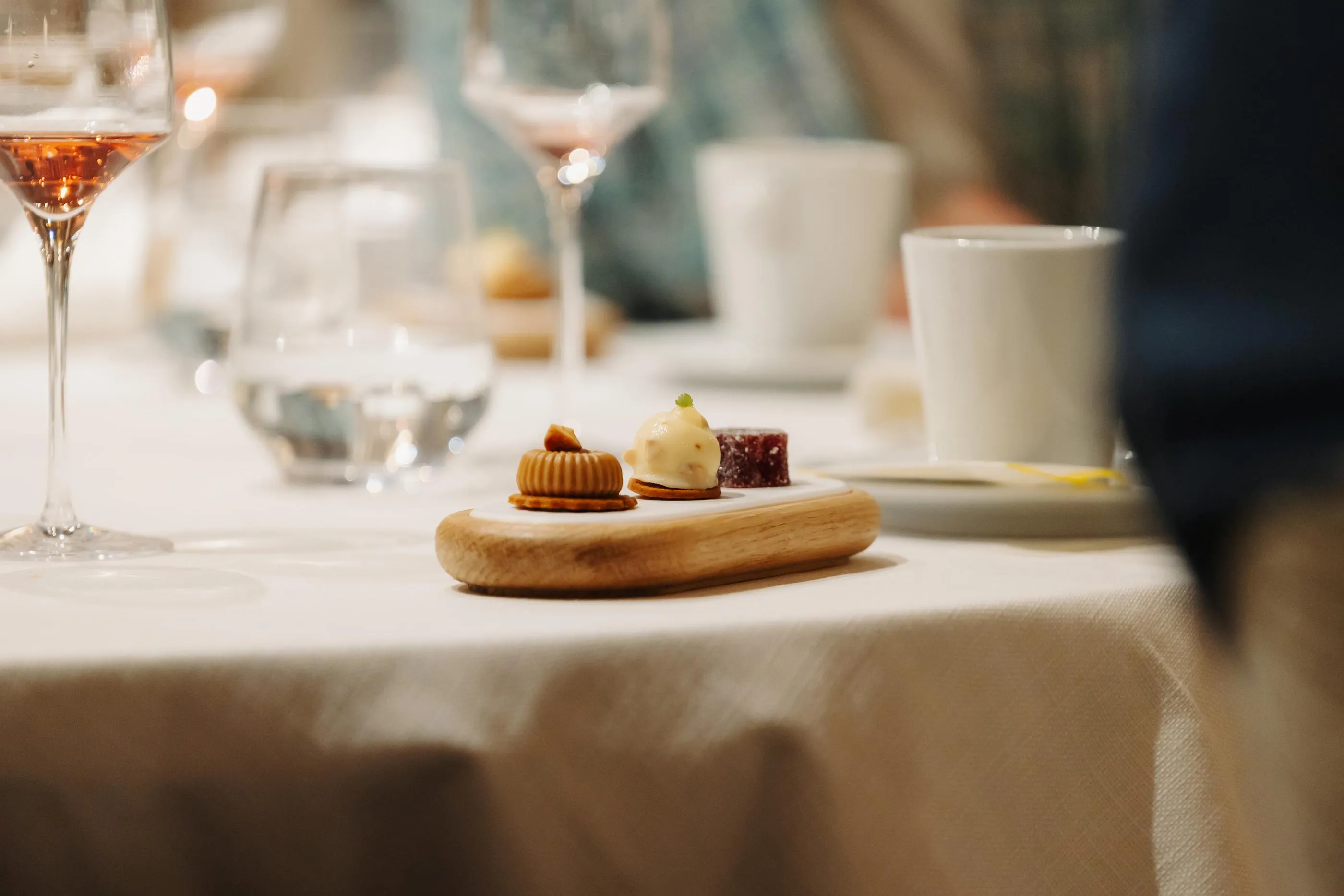 Kleine Holzschale mit drei Desserts und Weingläsern im Hintergrund auf einem mit einer weißen Tischdecke bedeckten Tisch