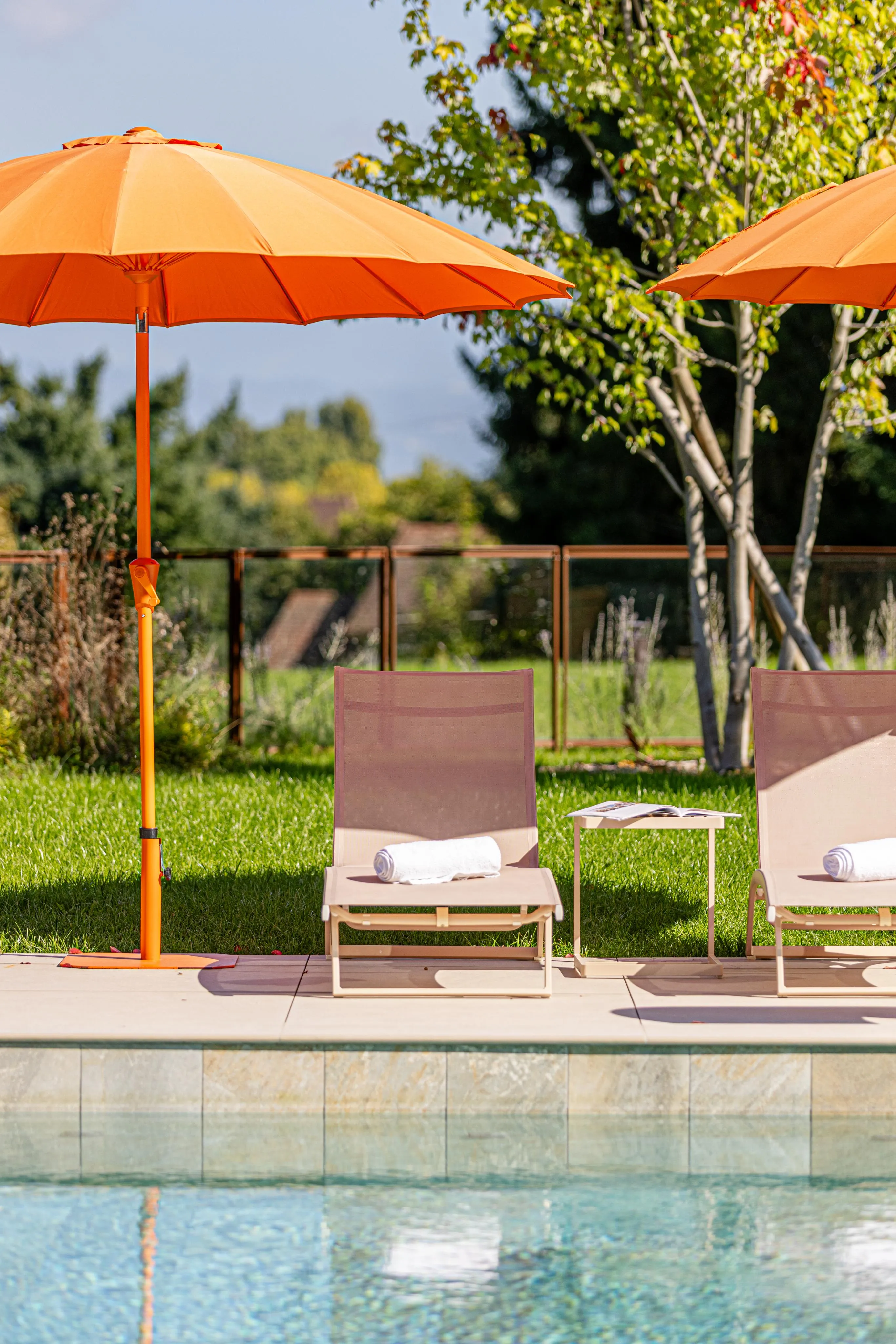 Deux transats avec serviettes et une table basse entre eux, sous des parasols orange, près d'une piscine avec un jardin en arrière-plan