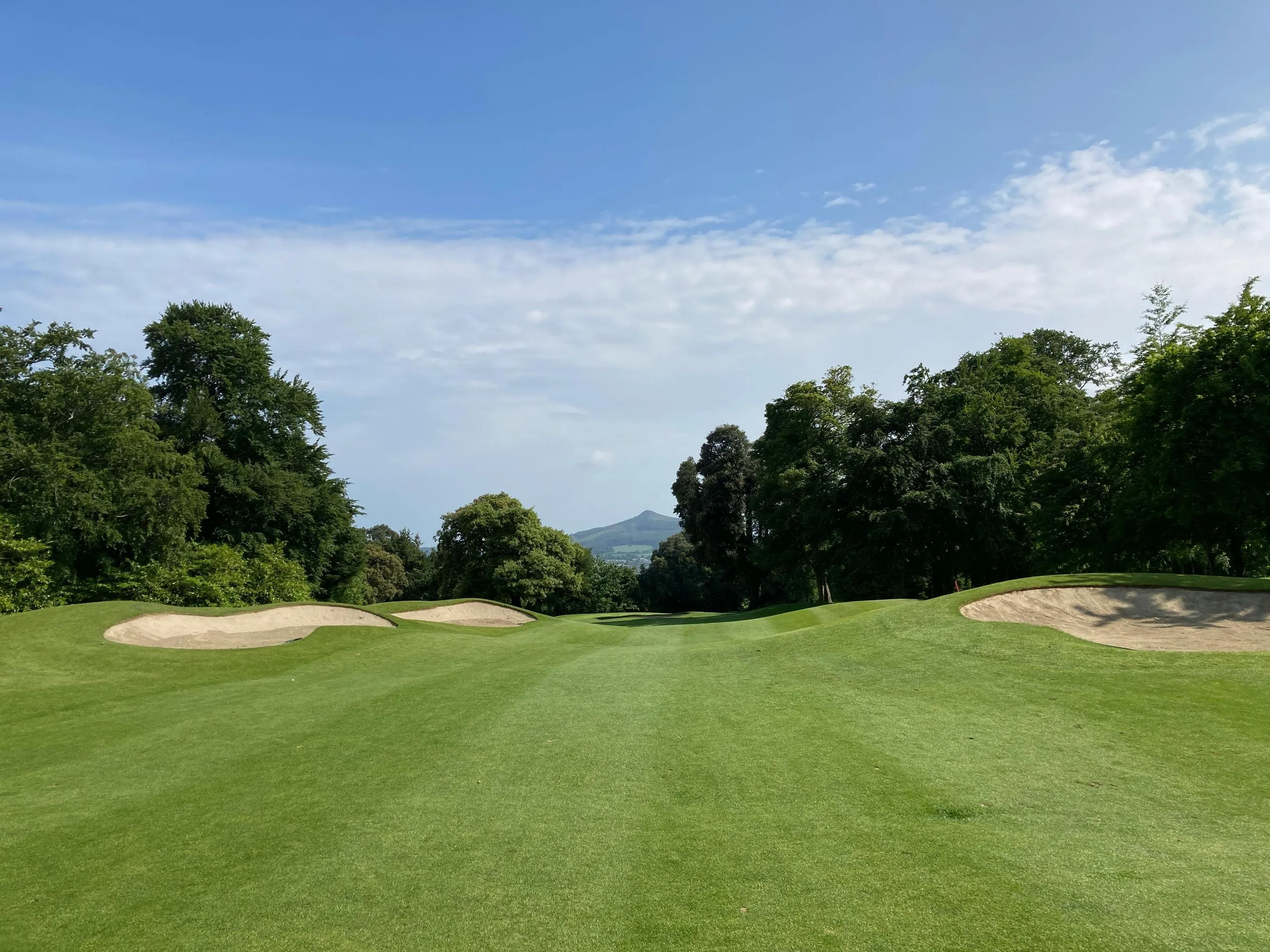 Un parcours de golf avec des bunkers de sable, entouré d'arbres et de collines sous un ciel partiellement nuageux