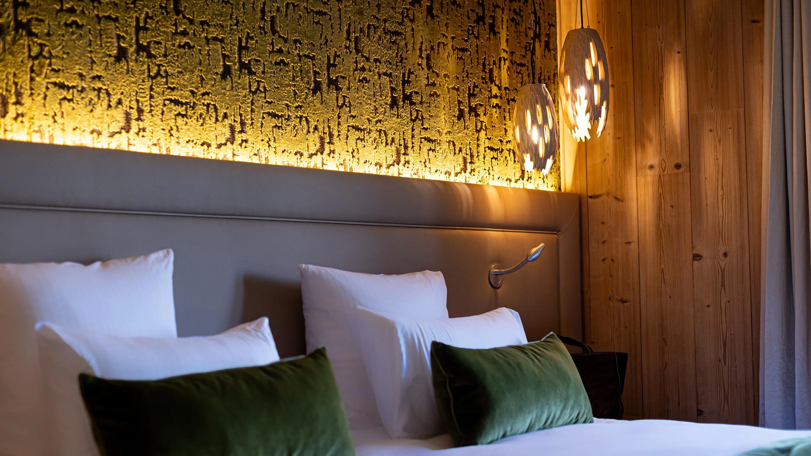 Bett mit grauer Stoffkopflehne, weißen und grünen Kissen, Wandlampen und einer mit einem goldenen Lichtstreifen beleuchteten Holzwand