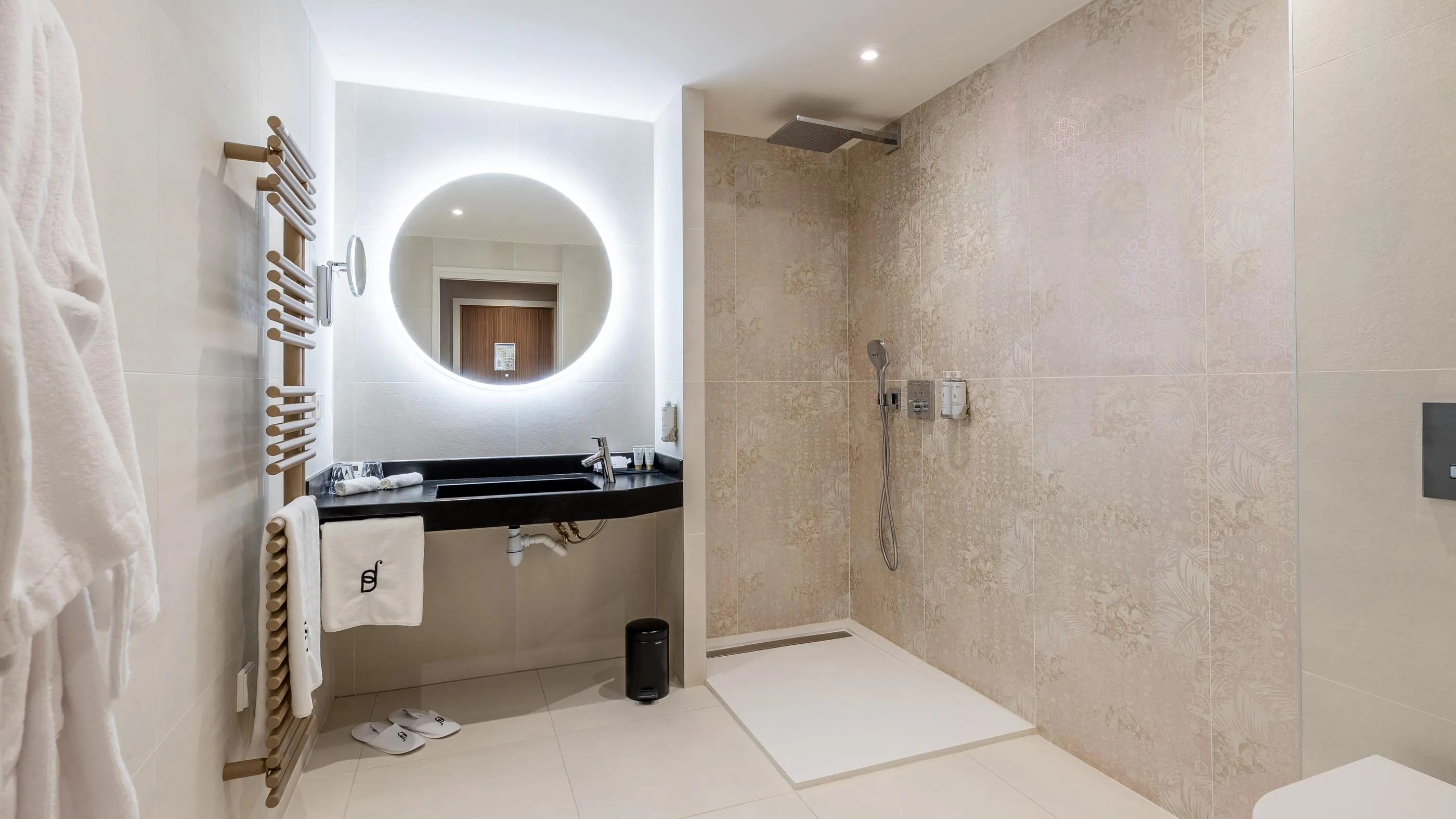 Moderne Badezimmer mit Waschbecken, beleuchtetem Rundspiegel, bodengleicher Dusche und aufgehängten Handtüchern