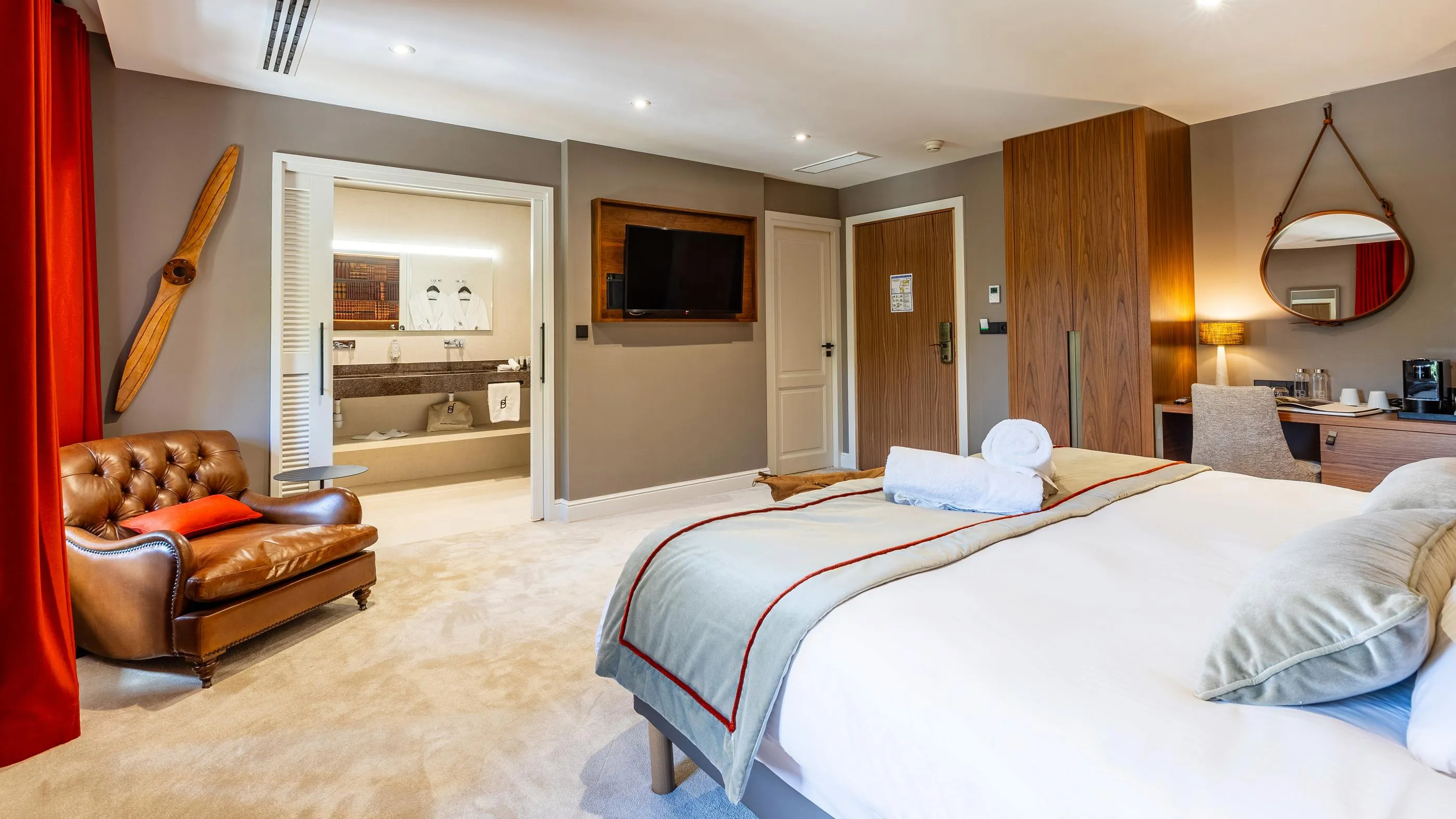 Hotelzimmer mit Doppelbett, Ledersessel, Fernseher, Spiegel und im Hintergrund sichtbarer Badezimmer
