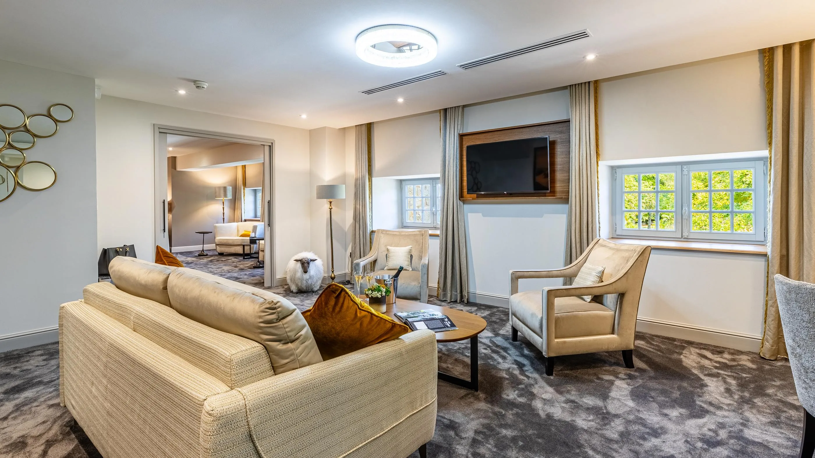 Wohnzimmer mit beigem Sofa, Sesseln, Couchtisch und Fernseher, Blick auf einen weiteren Raum mit Bett