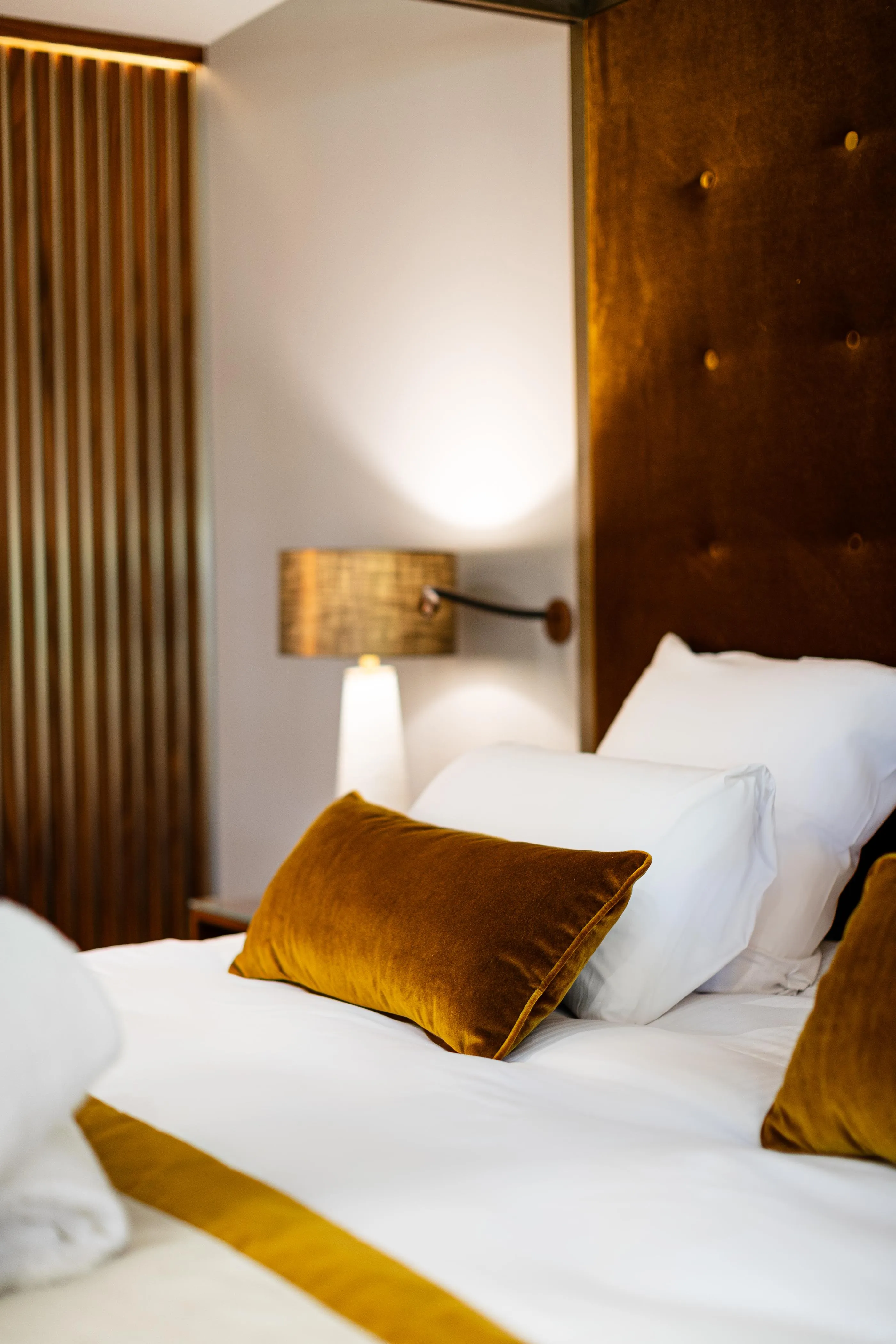 Bett mit weißen Laken, goldenen Samtkissen und einer eingeschalteten Nachttischlampe