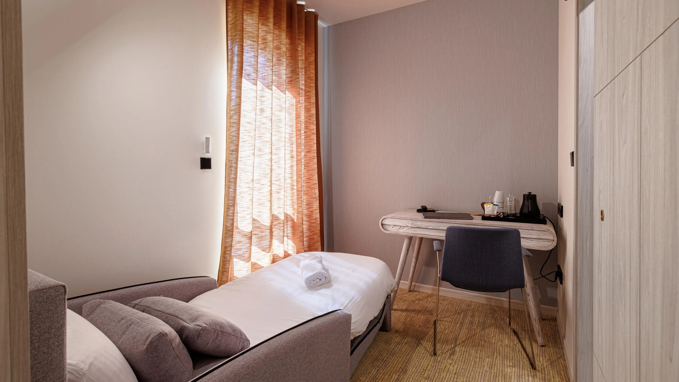Zimmer mit einem Einzelbett, einem Nachttisch, einem Fenster mit Vorhängen und einer strukturierten Wand
