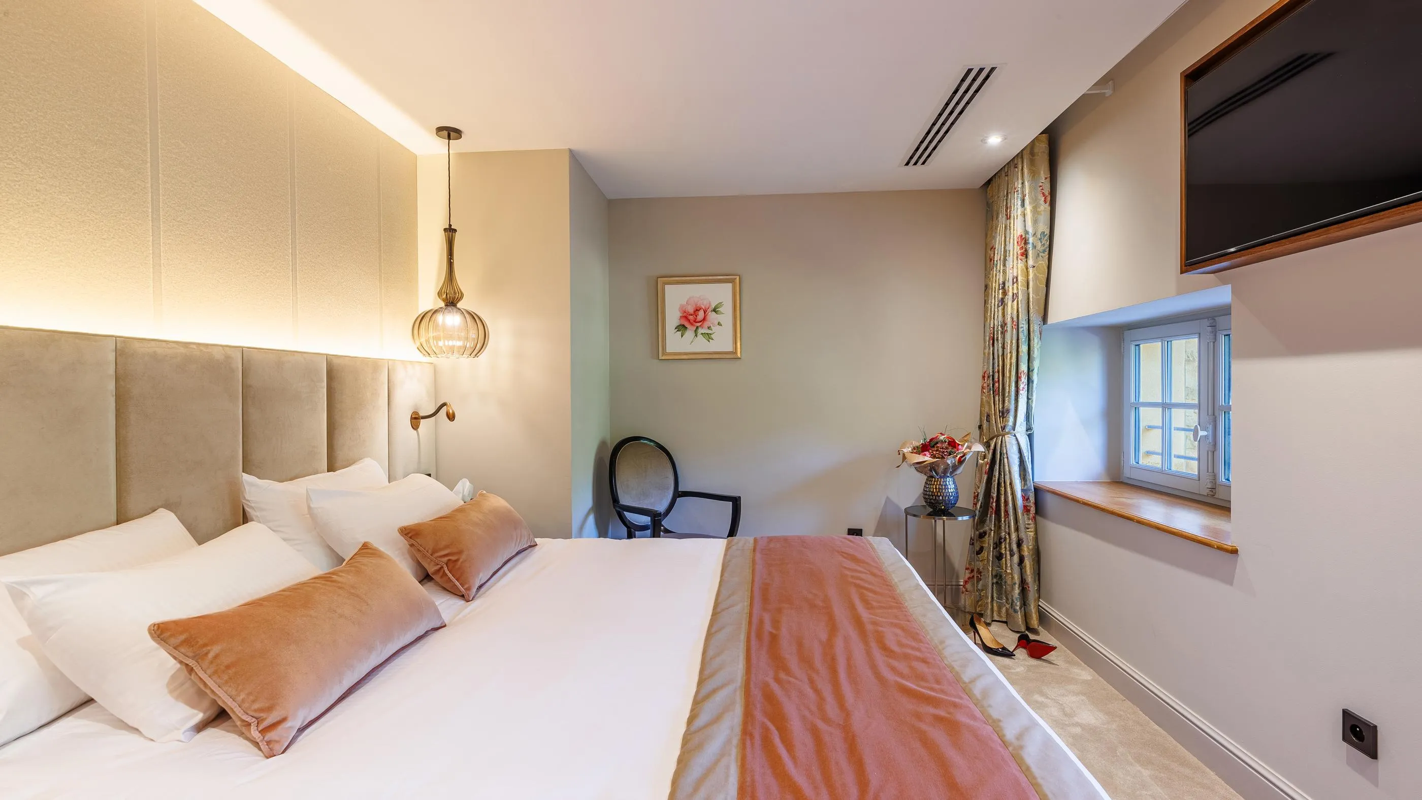 Chambre avec un lit double, des coussins peach, une lampe suspendue, un tableau floral et une fenêtre avec des rideaux