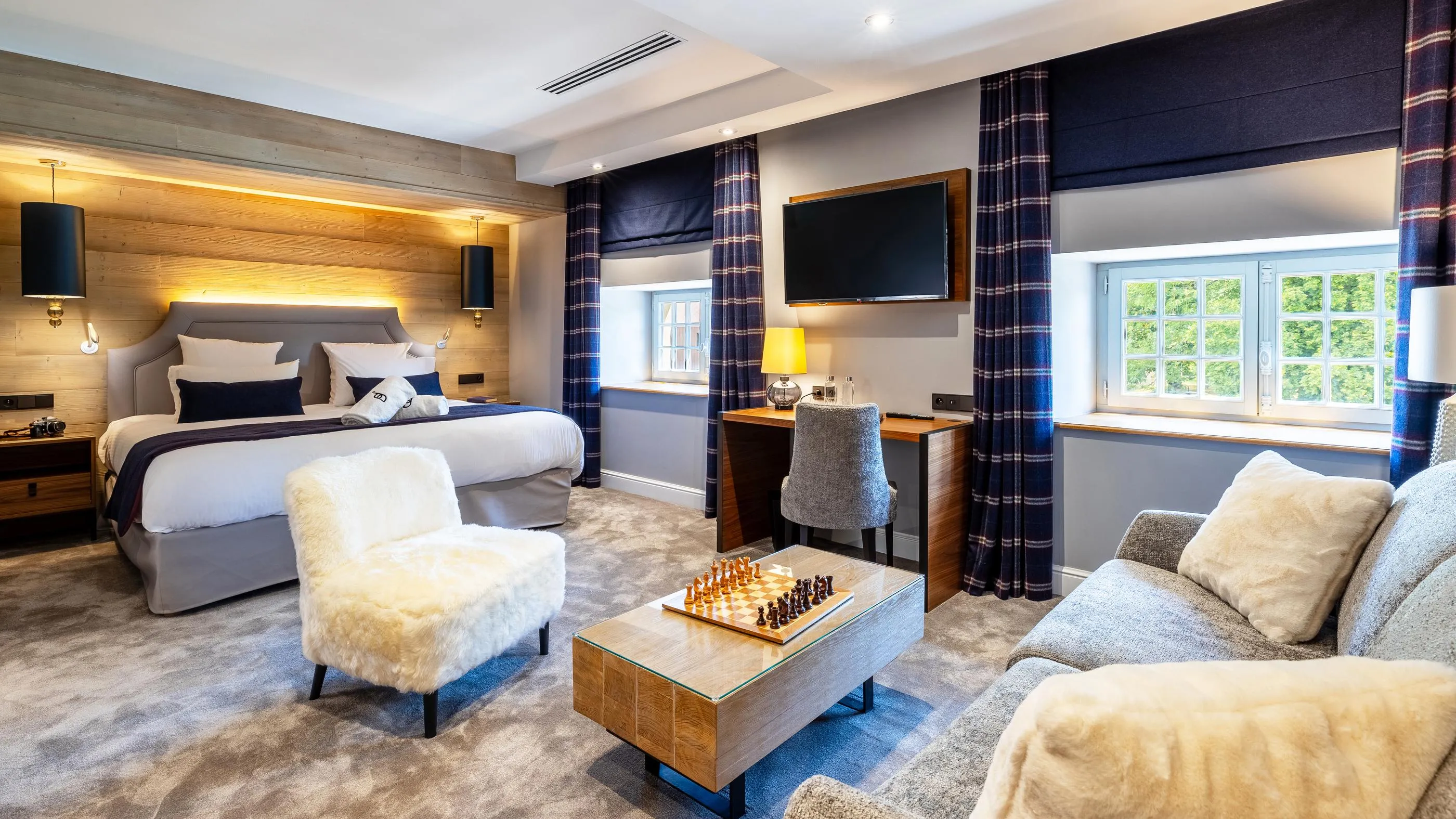 Chambre spacieuse avec lit, canapé, fauteuil, table basse avec échiquier, bureau et télévision, décorée dans des tons gris et bleu avec des éléments en bois et des fenêtres