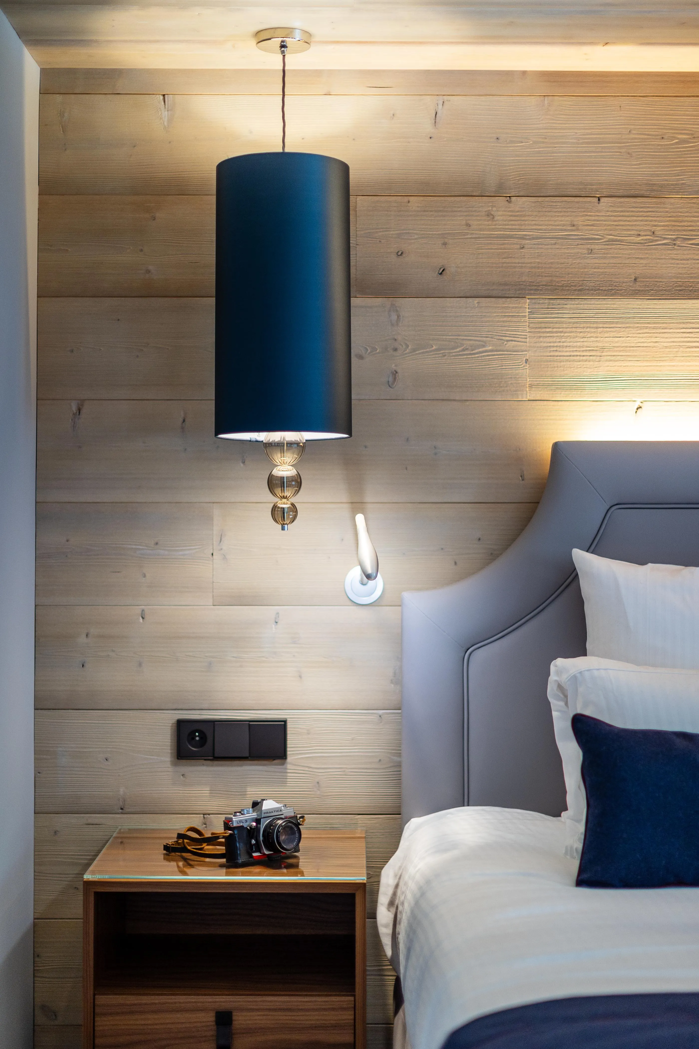 Une chambre avec un lit, une lampe suspendue, une table de chevet avec un appareil photo et une lampe murale