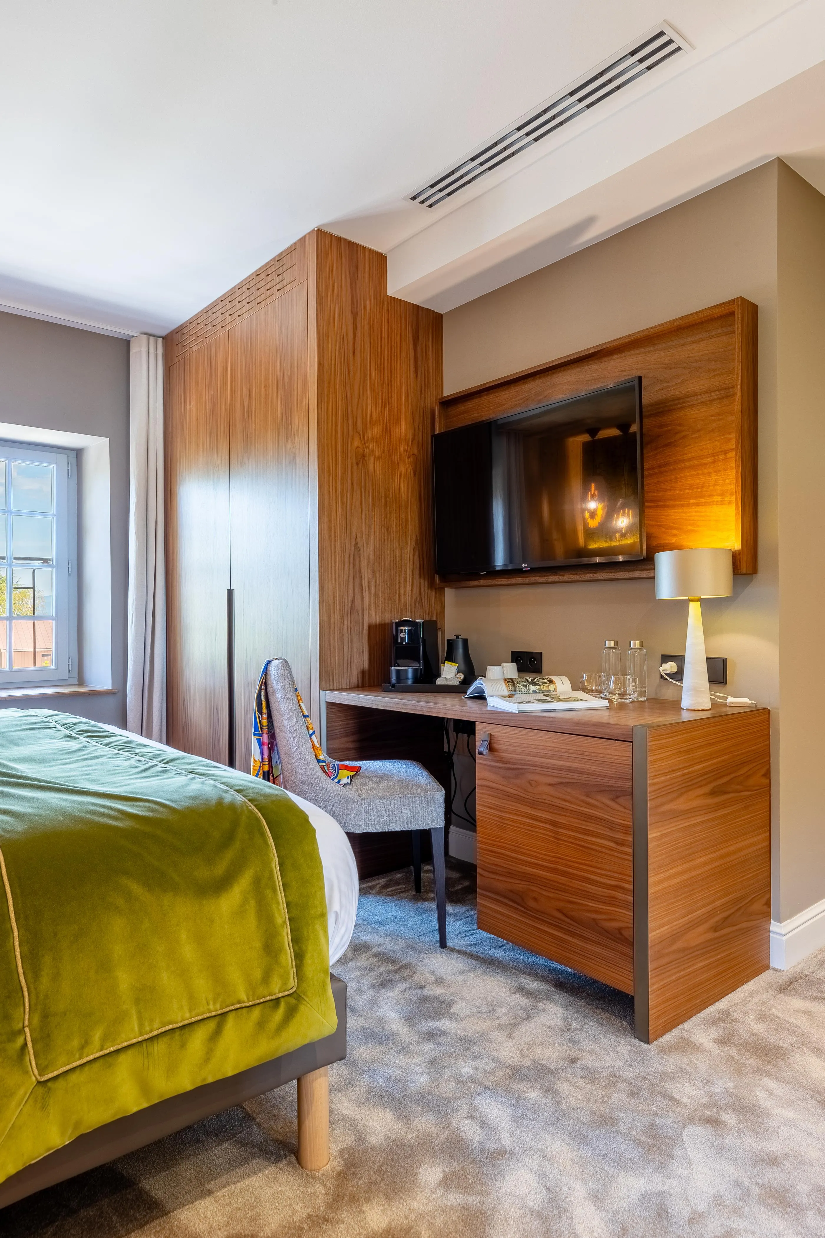 Zimmer mit einem Bett links, das mit einer grünen Decke bedeckt ist, einem Holznachttisch mit einer Lampe, einem eingeschalteten Fernsehbildschirm und einem Sessel am Fenster