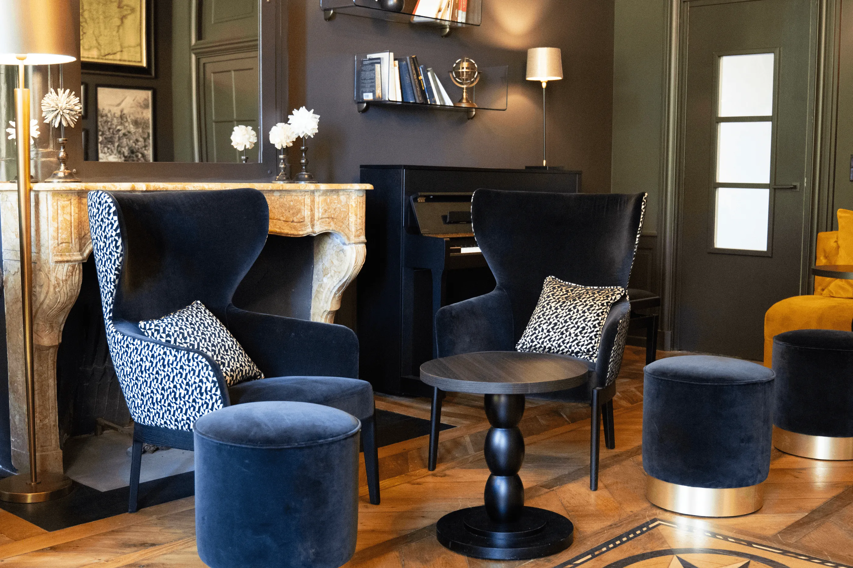 Salon mit Sesseln und Ottomanen aus Samt, einem Kamin und Regalen mit Büchern und Blumen