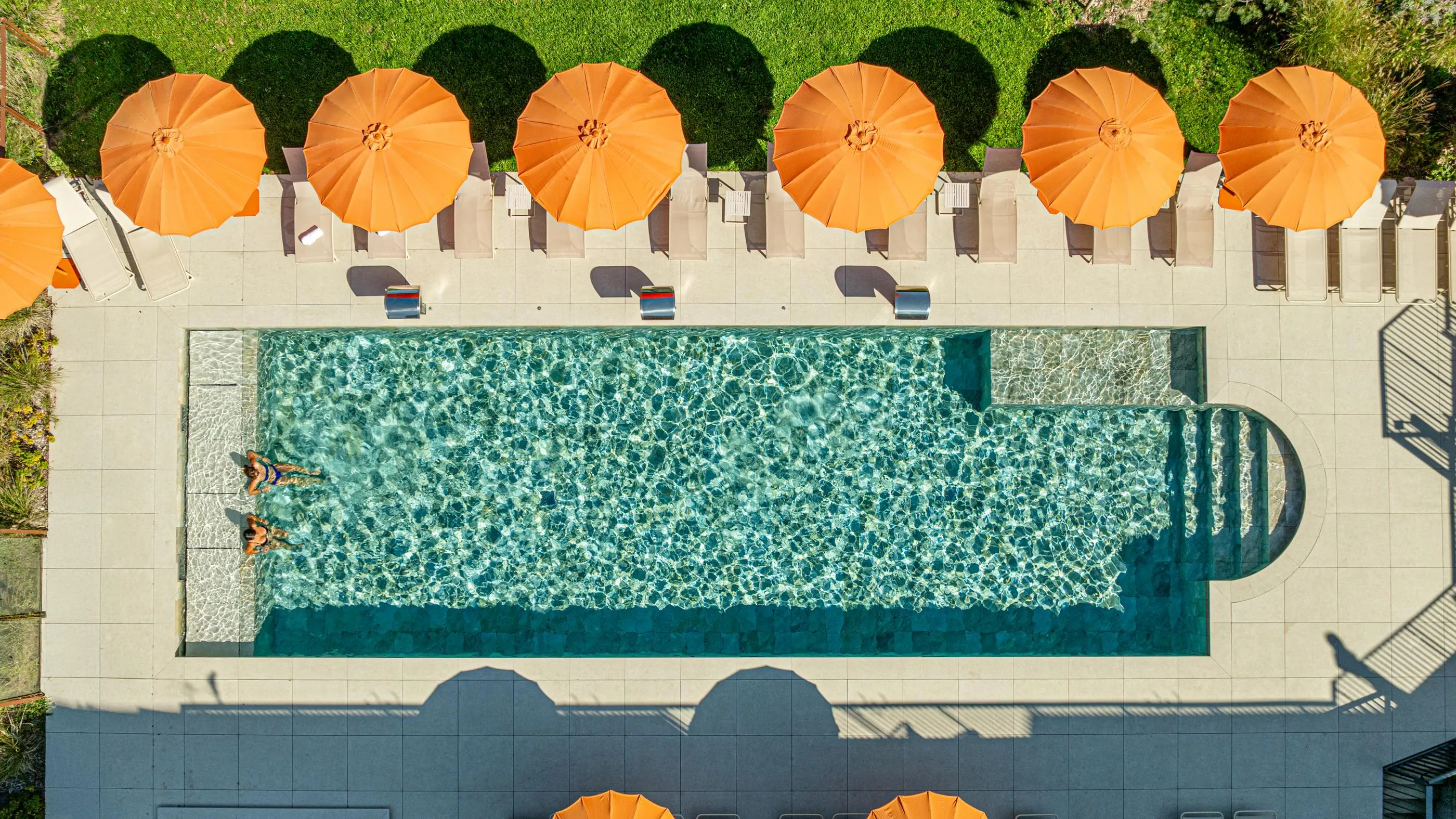 Vue aérienne d'une piscine rectangulaire avec deux personnes nageant, entourée de transats et de parasols orange