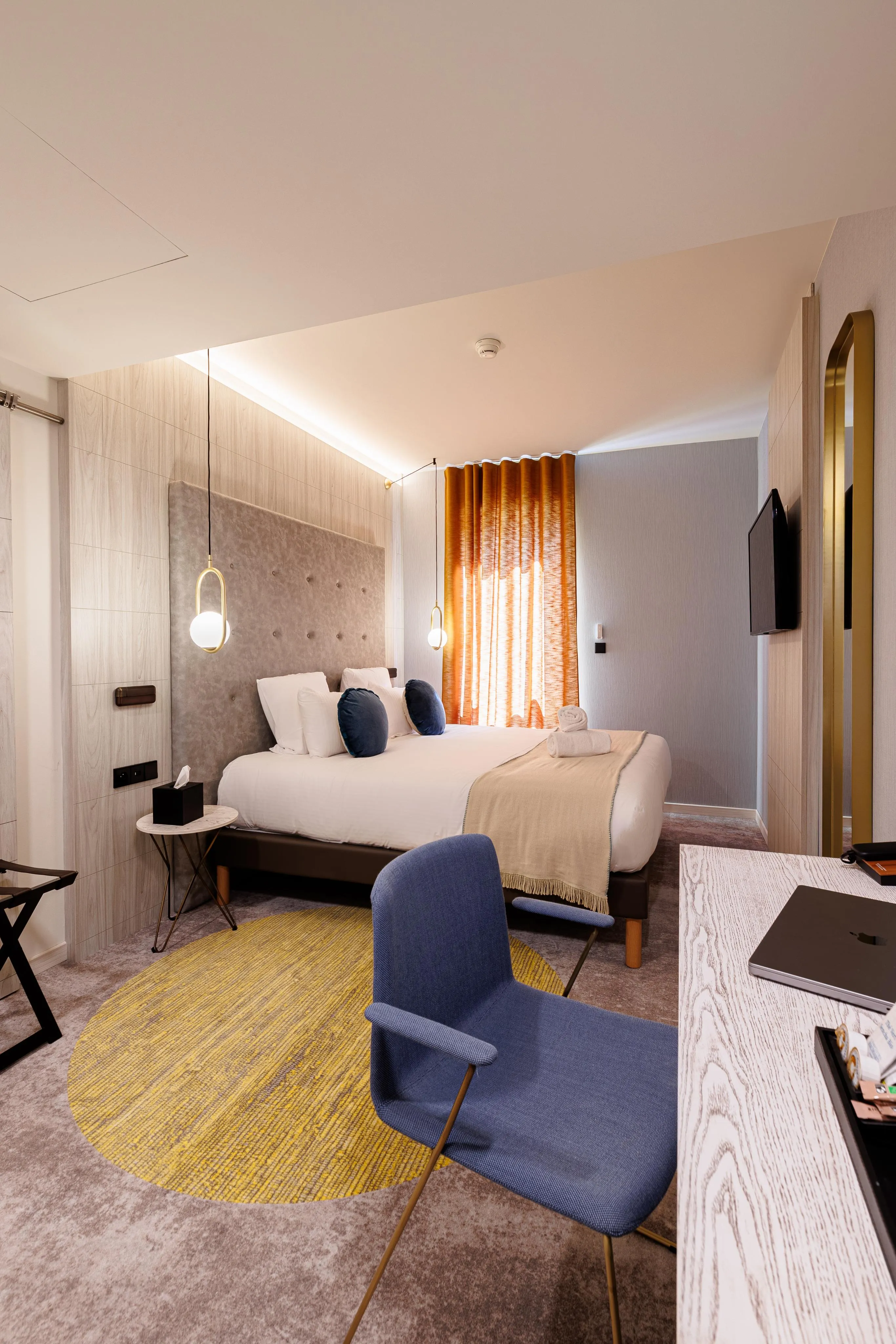 Chambre d'hôtel avec un lit double, une chaise bleue, un bureau, des lumières murales et des rideaux orange