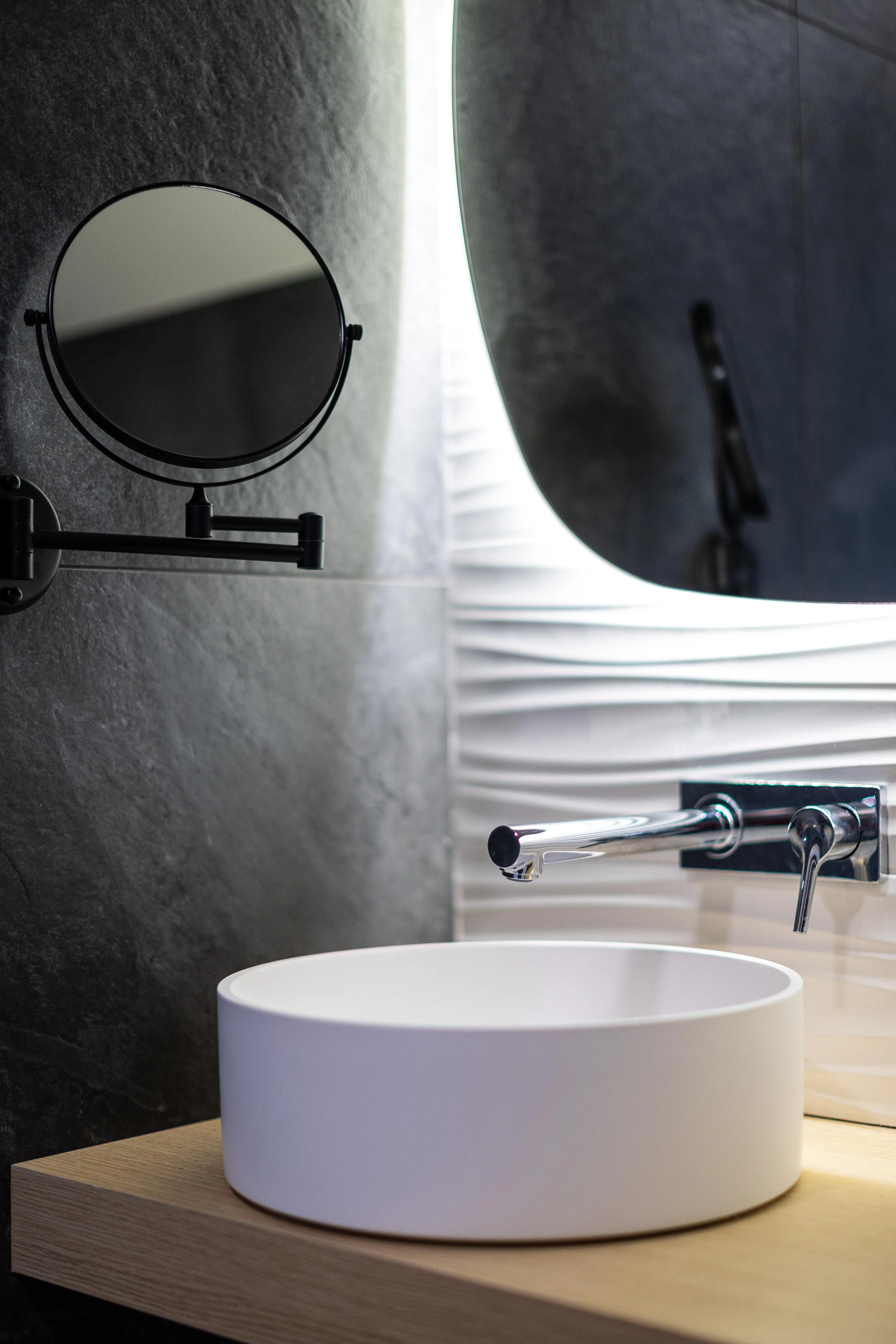 Un lavabo blanc sur un meuble en bois avec un robinet en métal et un miroir rond à bras articulé sur le mur gris