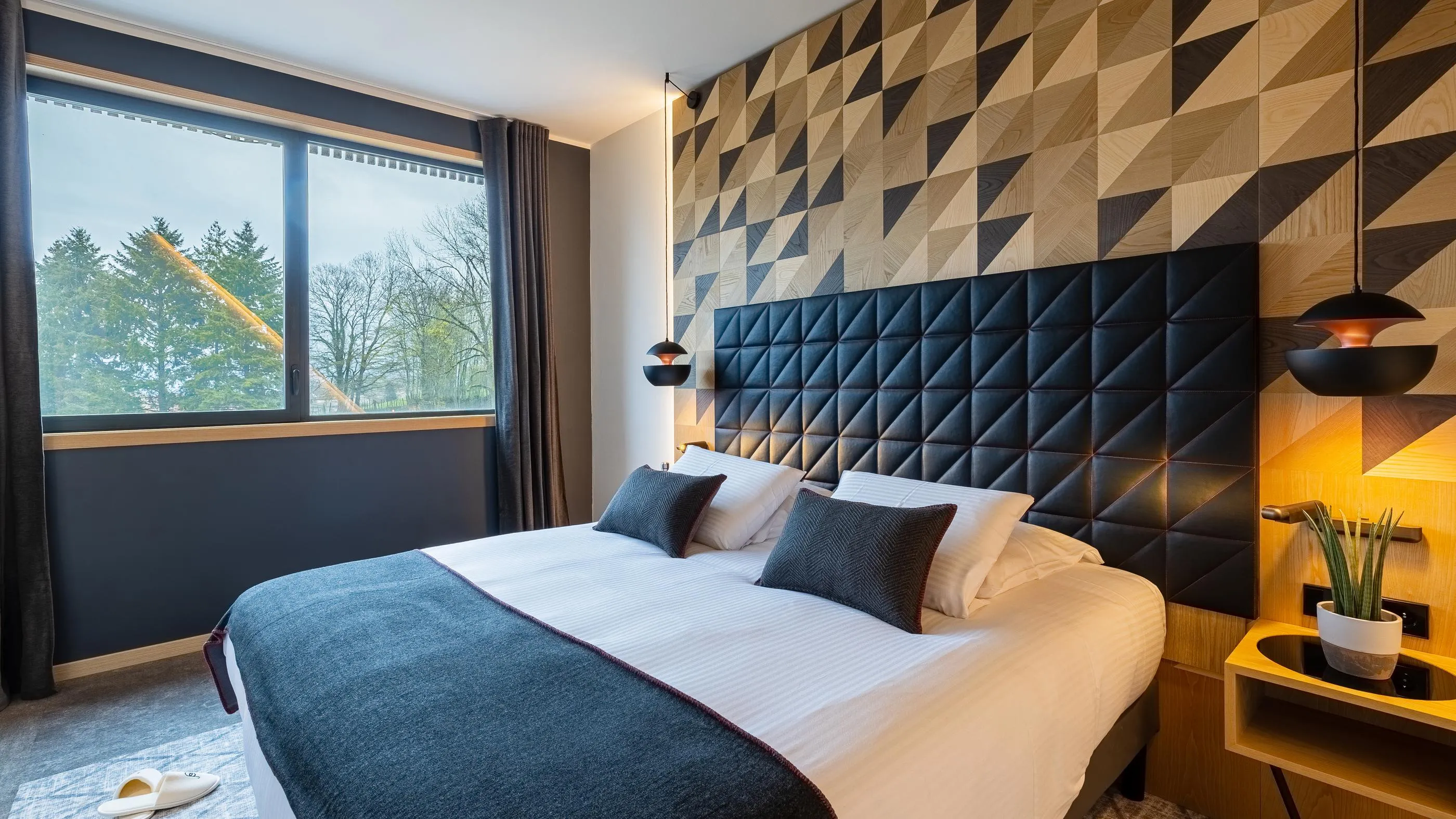 Chambre avec un lit double blanc et gris, une tête de lit en bois géométrique, des lampes noires et une vue sur des arbres par une grande fenêtre