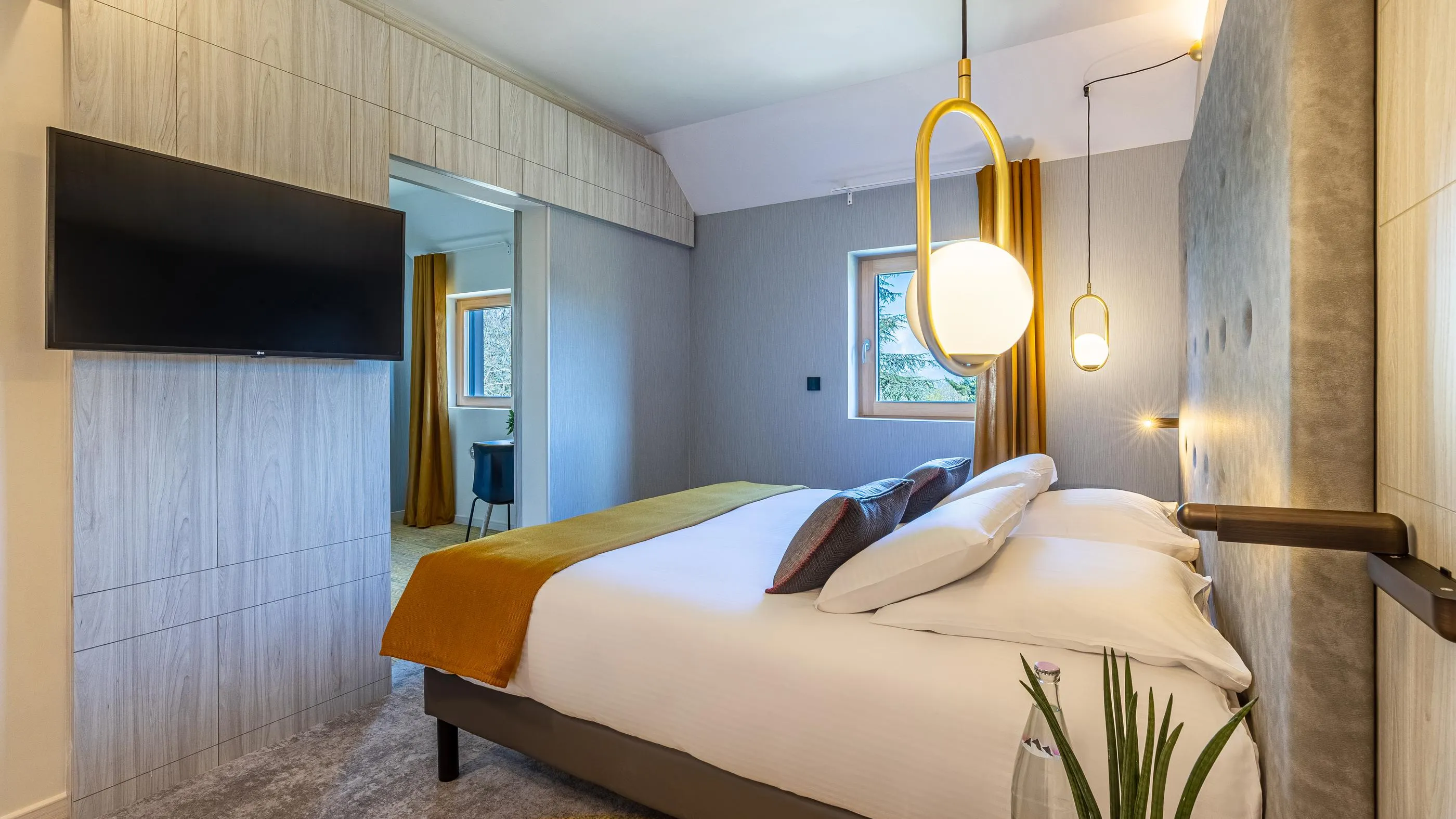 Chambre moderne avec lit, télévision, lampes suspendues et vue sur une autre pièce par une porte ouverte