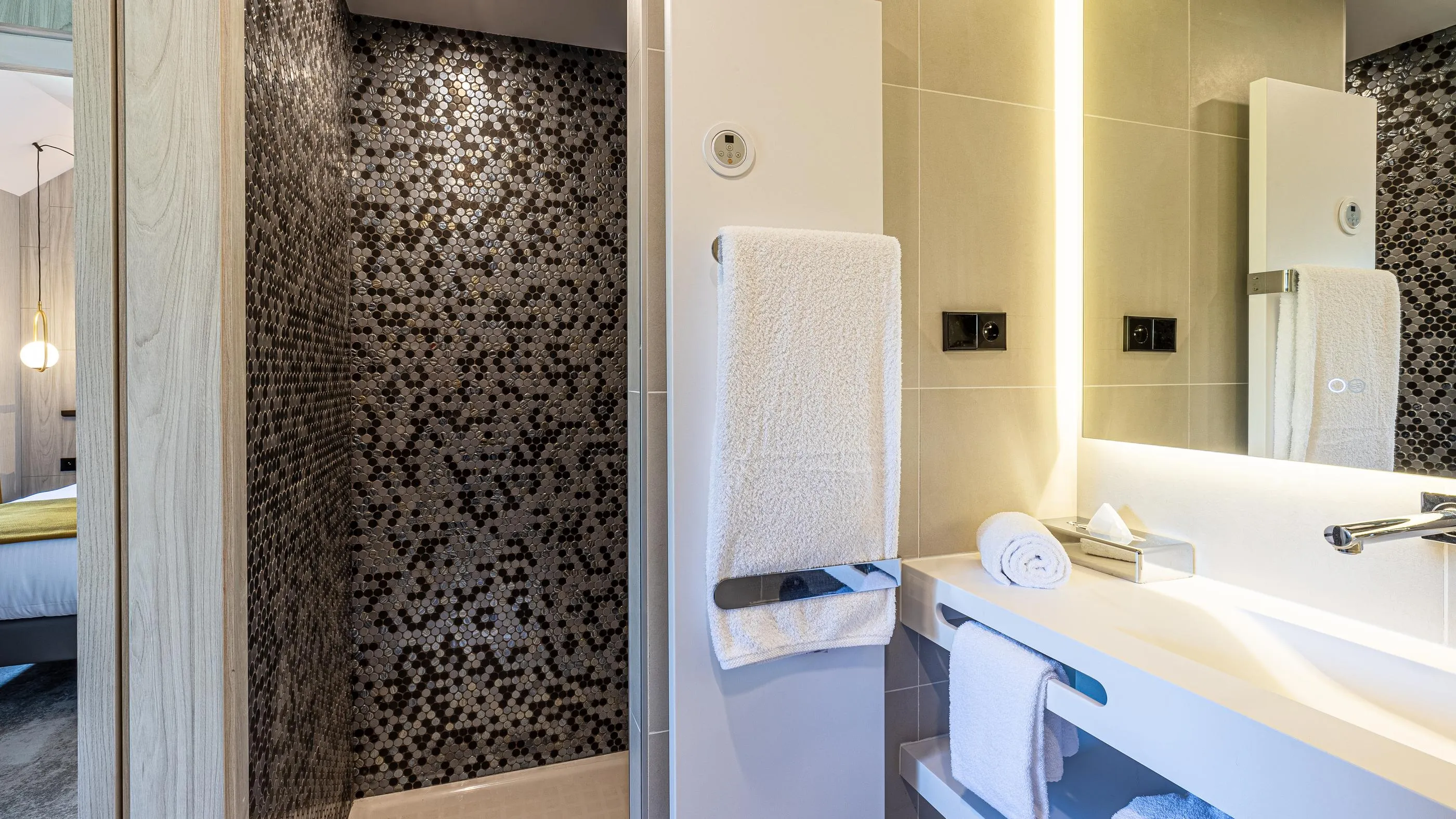 Salle de bain moderne avec douche à mosaïque, lavabo, serviettes et miroir reflétant une partie de la chambre
