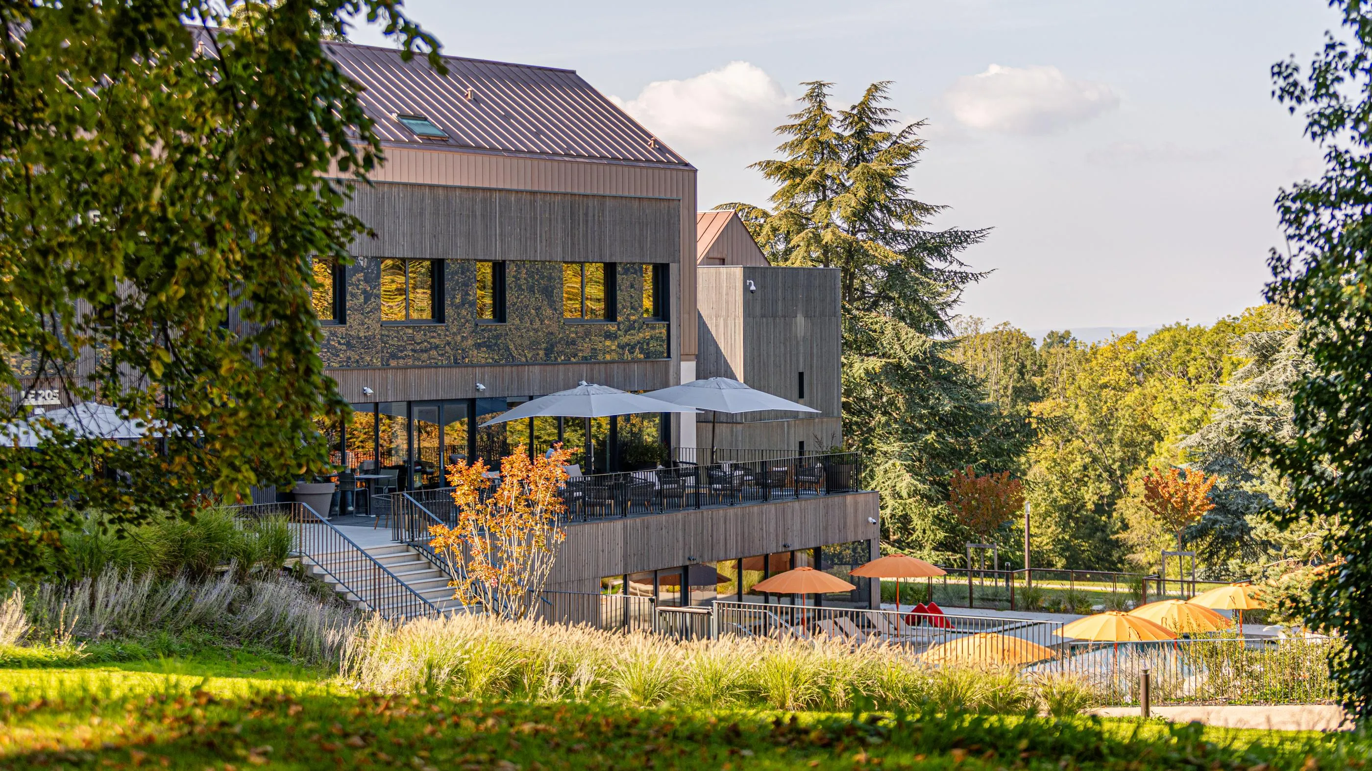 Bâtiment moderne avec terrasse et parasols entouré d'arbres et de végétation en automne