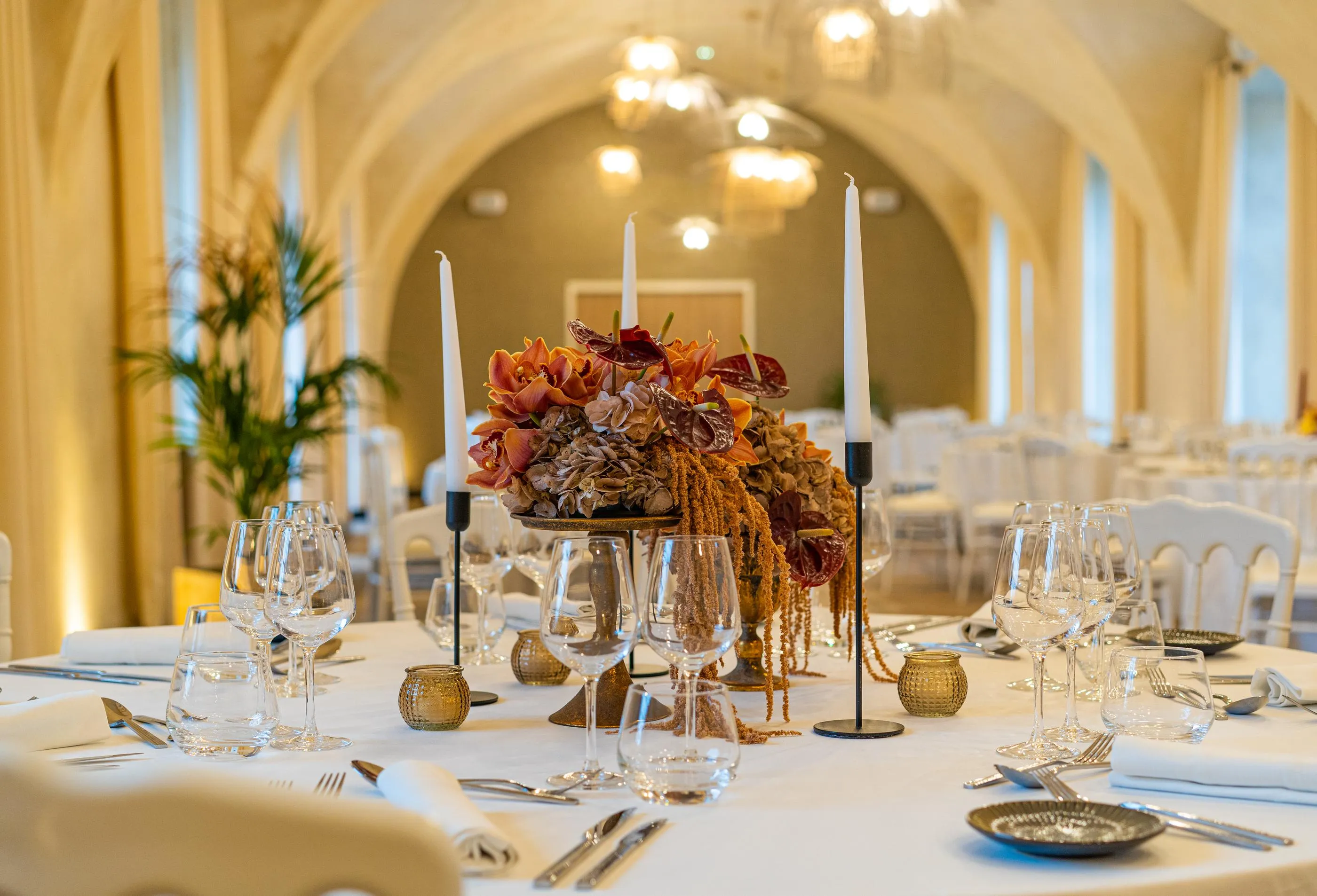 Table dressée avec verres, couverts, bougies et un centre de table floral dans une salle aux arches éclairées
